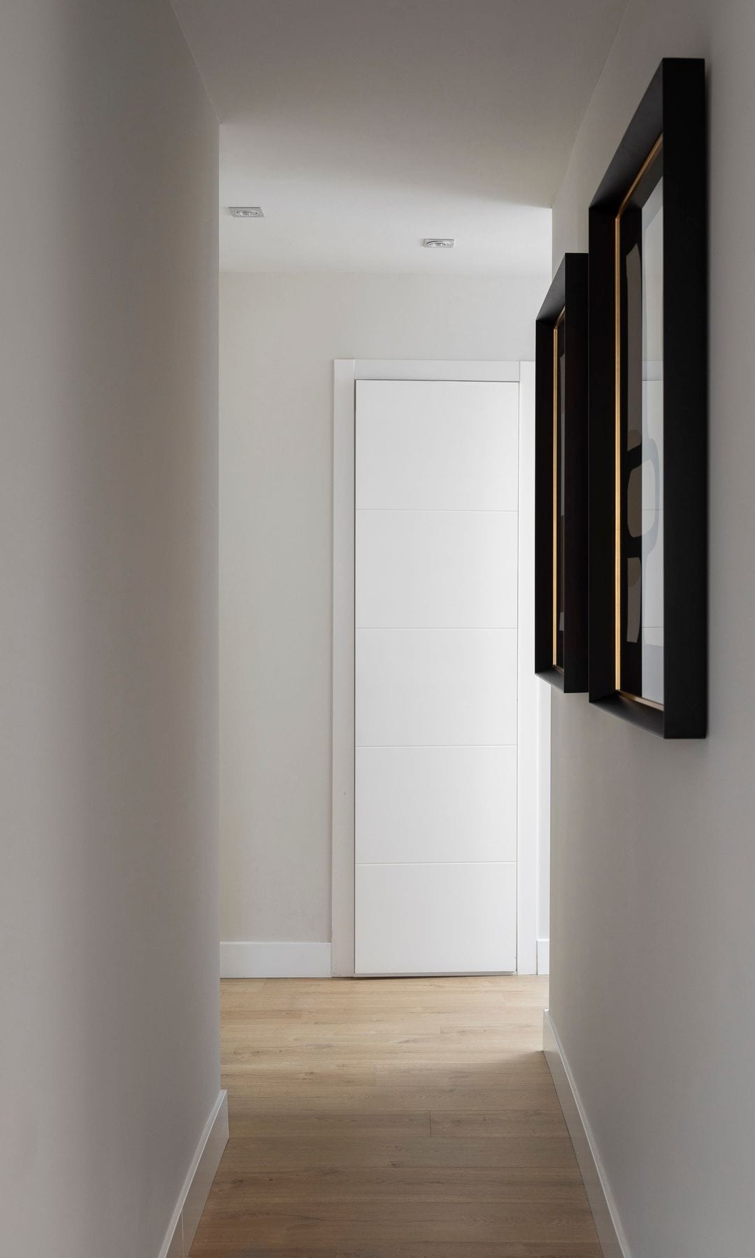 Pasillo blanco con cuadros con el marco negro, focos empotrados en el techo y suelo de madera