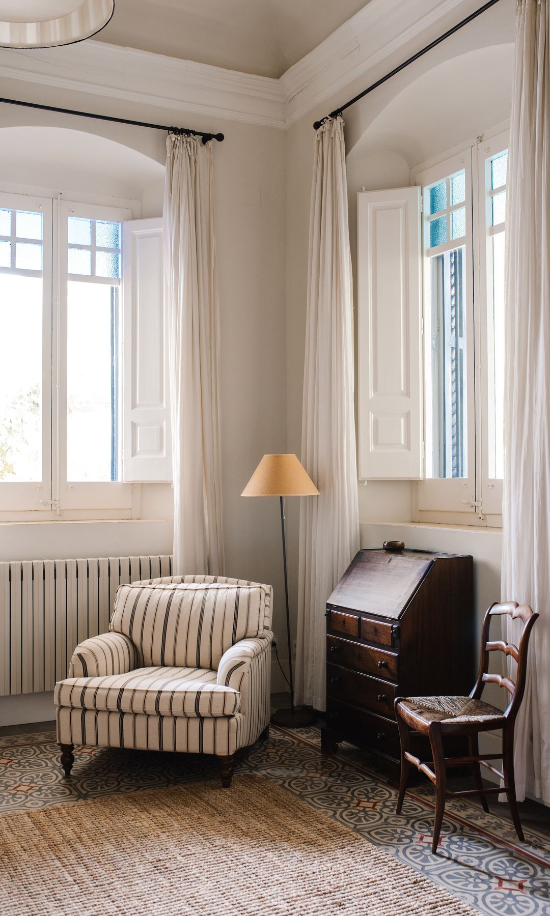 Rincón de lectura con sillón con tela de rayas, secreter antiguo, alfombra, lámpara de pie, silla de madera, visillos blancos 