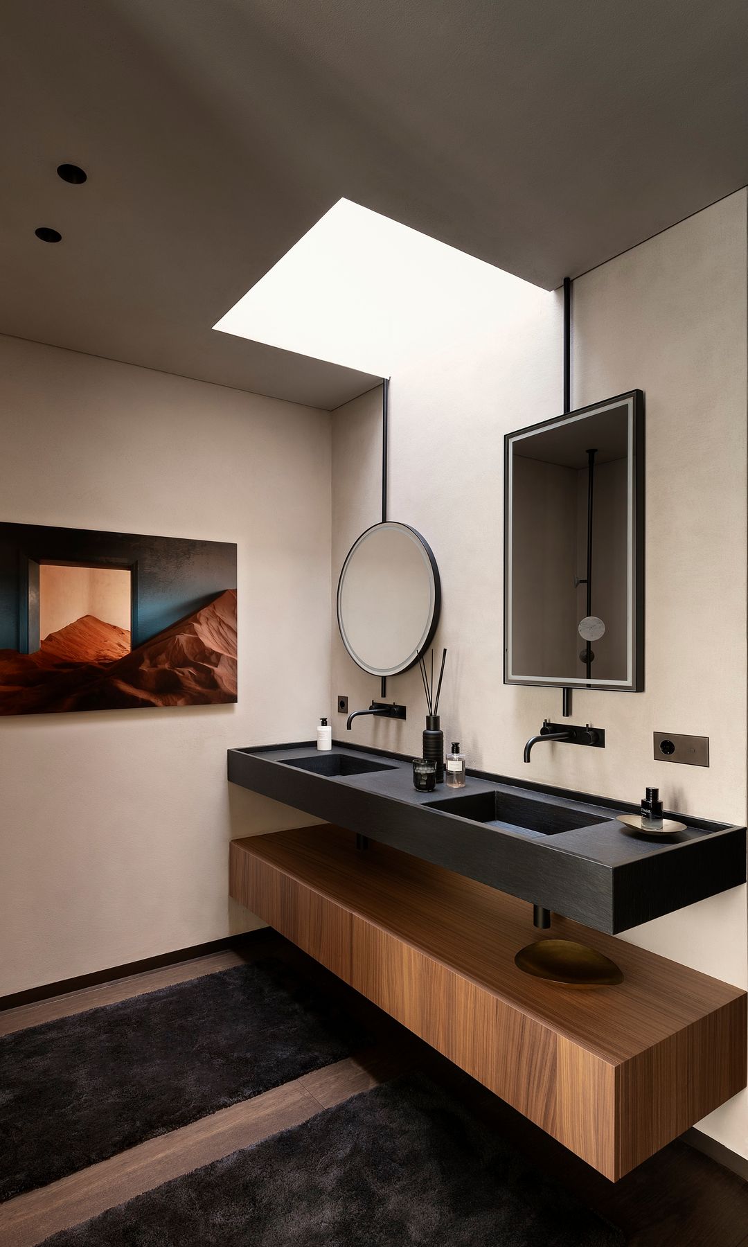 Baño con lavabo doble negro, mueble de madera, suelo de madera, ventana de techo, espejo redondo y espejo rectangular 
