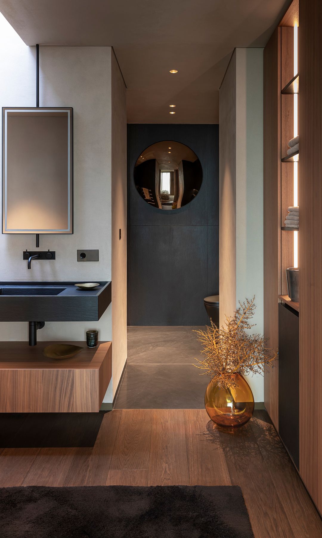 Baño con suelo de madera paredes, negras, estanterías y espejo efecto redondo
