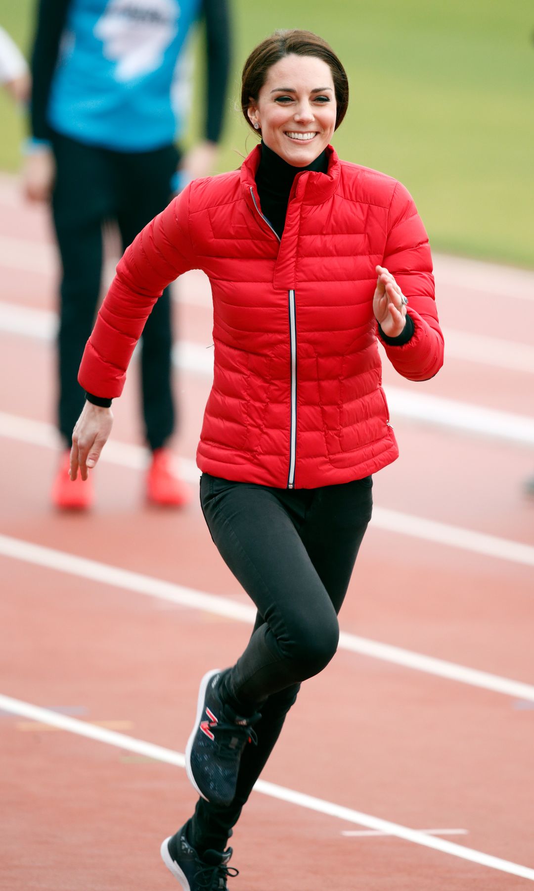 Kate Middleton corre sonriendo en una pista de atletismo, vestida con chaqueta roja y leggings negros.