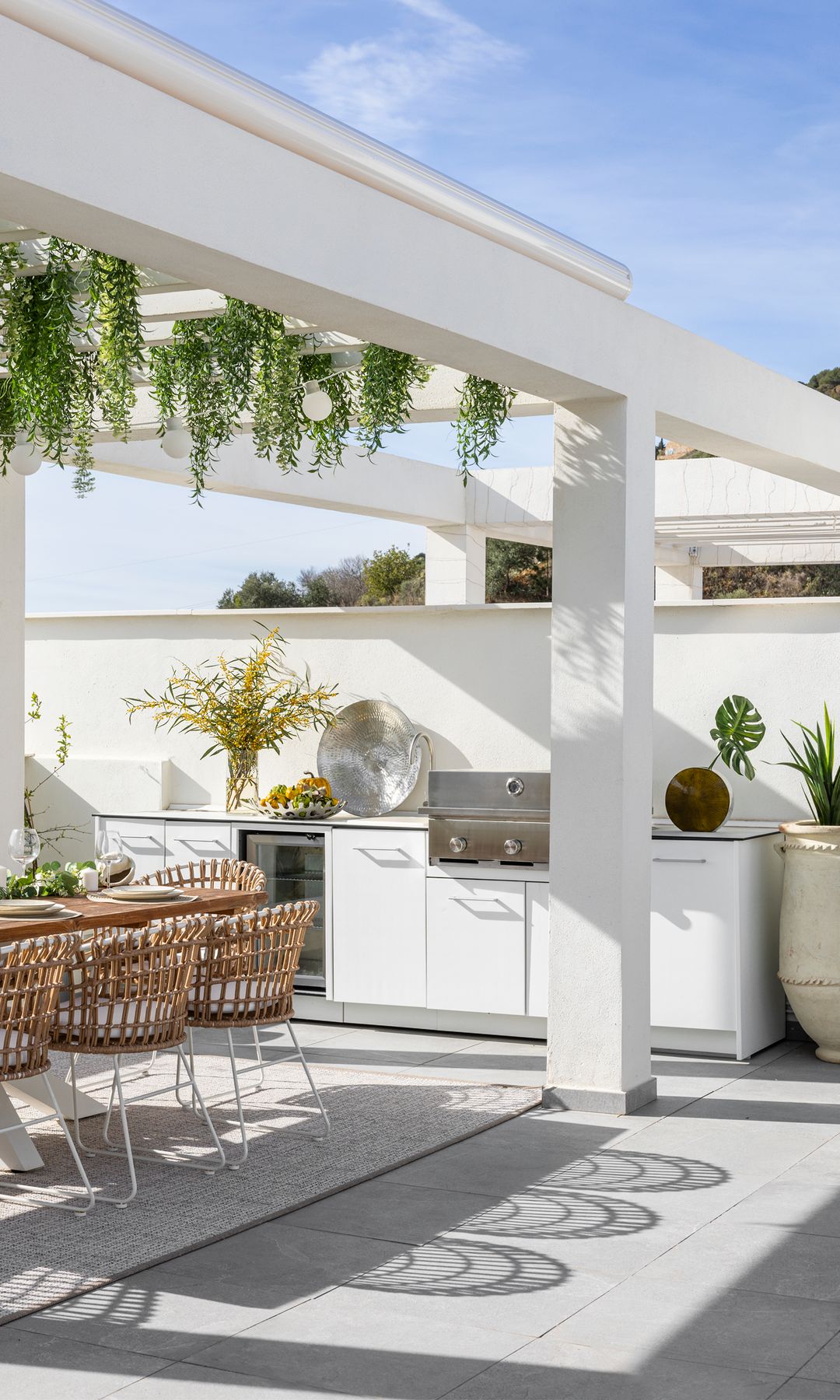 Terraza con cocina de exterior en blanco, bajo pérgola con mesa de comedor y sillas