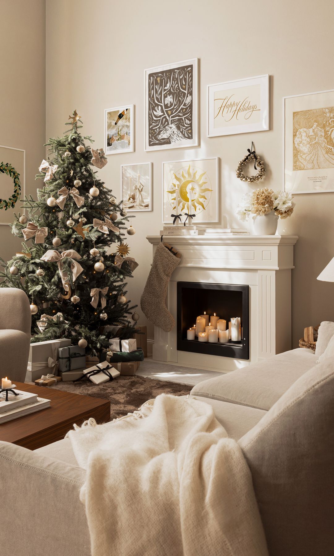 Salón con chimenea, árbol de Navidad, láminas en las paredes sobre la chimenea, velas en la chimenea, manta sobre el sofá, adornos dorados y blancos