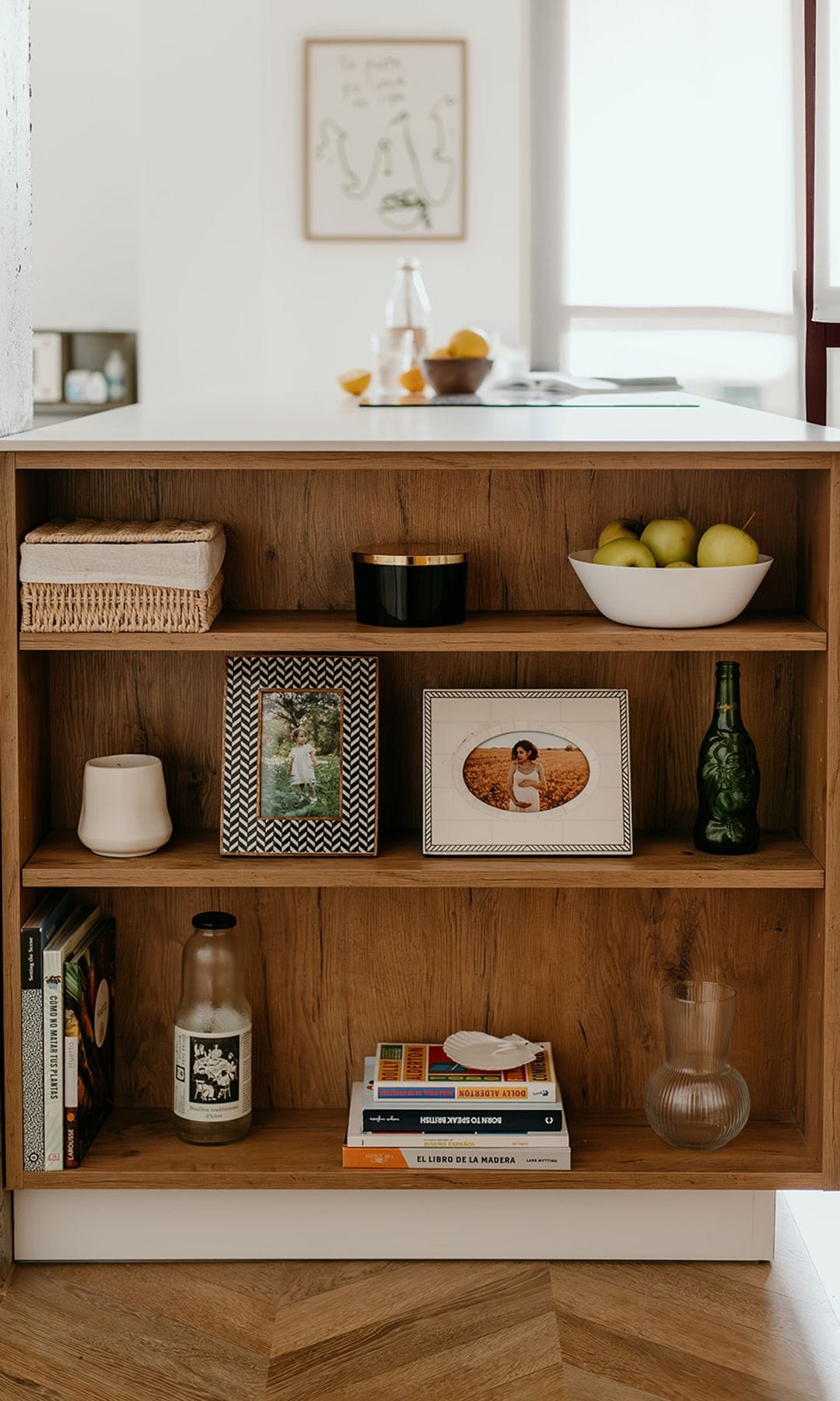 Estantería en un lateral de una isla, con baldas de madera, libros, fotografías, botellas y cestos, suelo de madera 