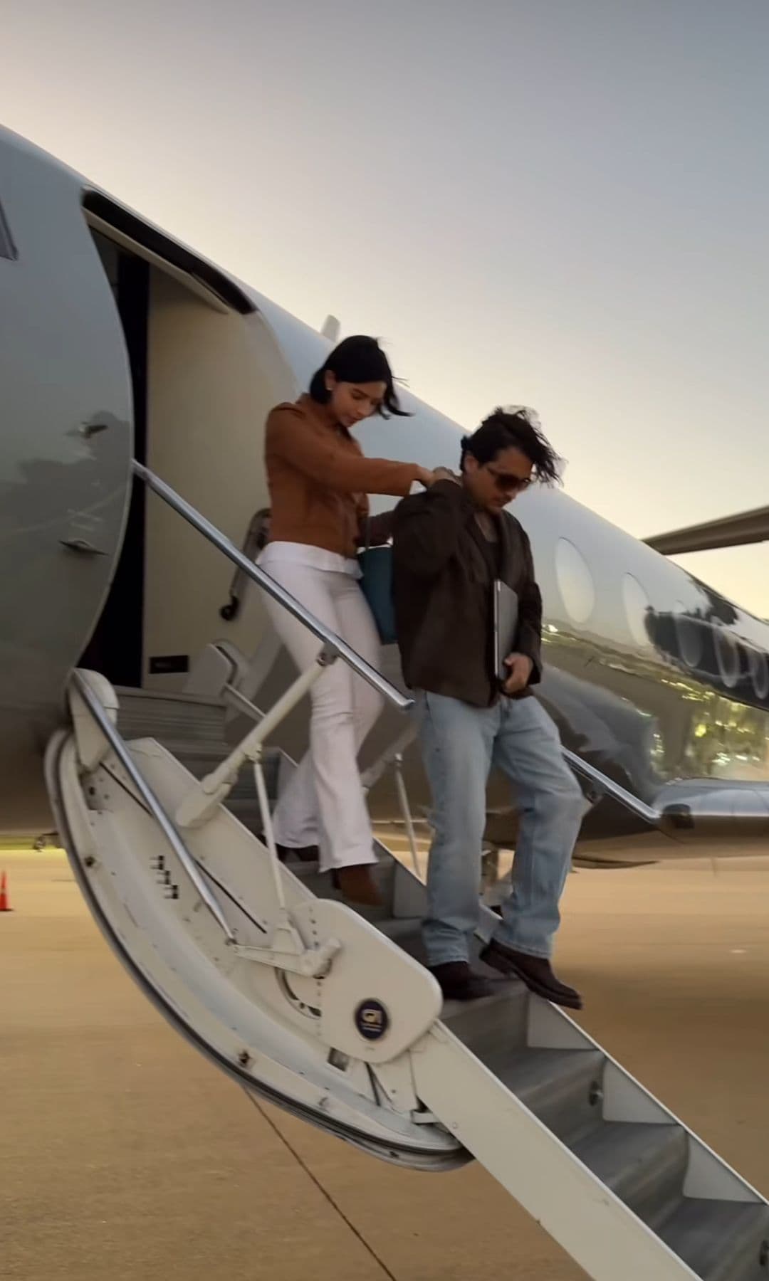 Ángela Aguilar y Christian Nodal descienden del avión; ella se apoya en él.