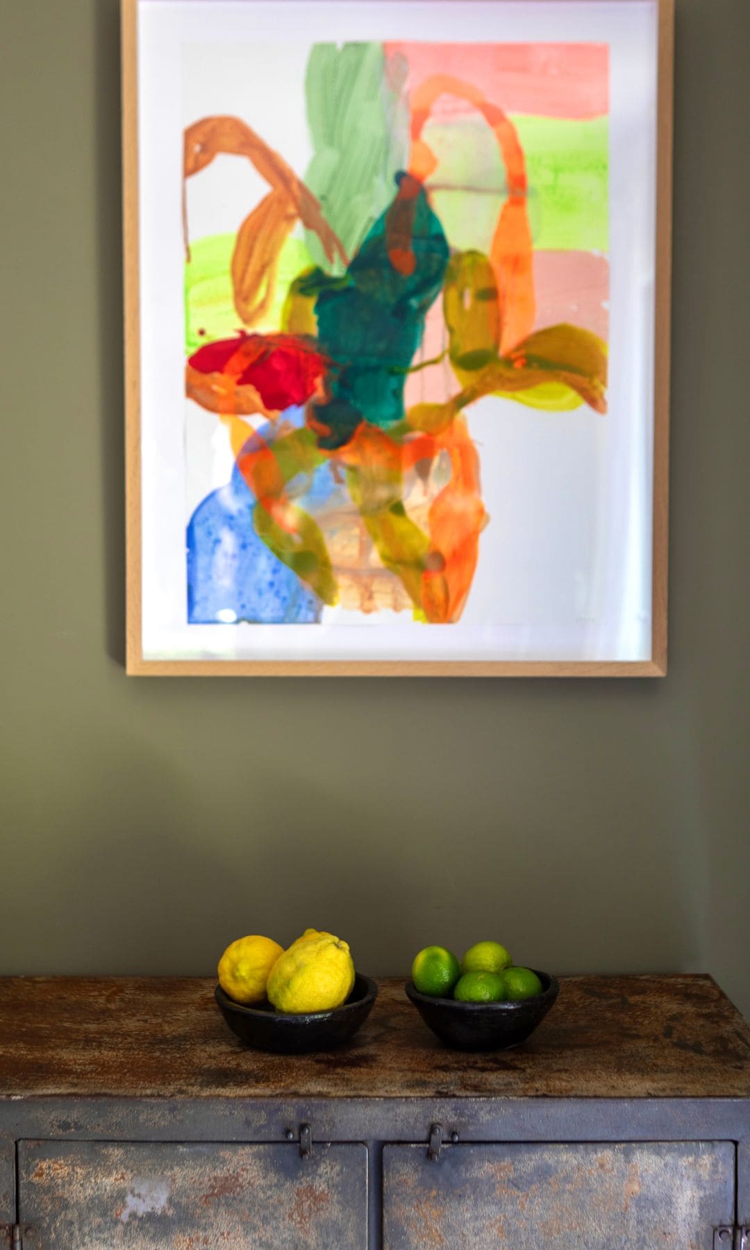 Mueble antiguo con fruteros negros con limones y limas, cuadro abstracto en naranjas, verdes y rojos