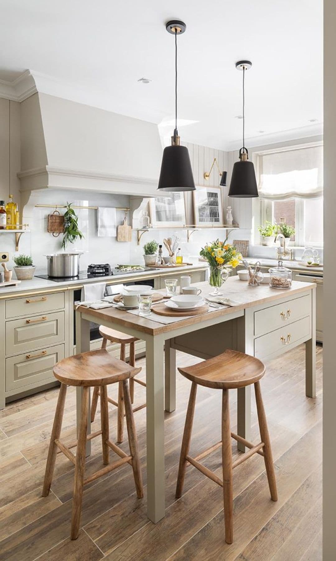 Cocina de estilo British con mesa alta en el centro con cajones y taburetes, suelo de madera, lámparas de techo negras
