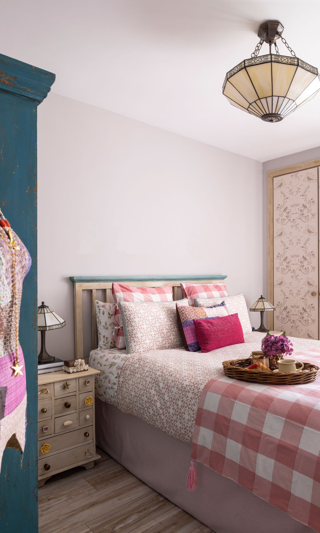 Dormitorio con armario con las puertas empapeladas, lámpara de techo, ropa de cama de cuadros rojos y blancos, cabecero de lamas de madera, maniquís en un rincón 