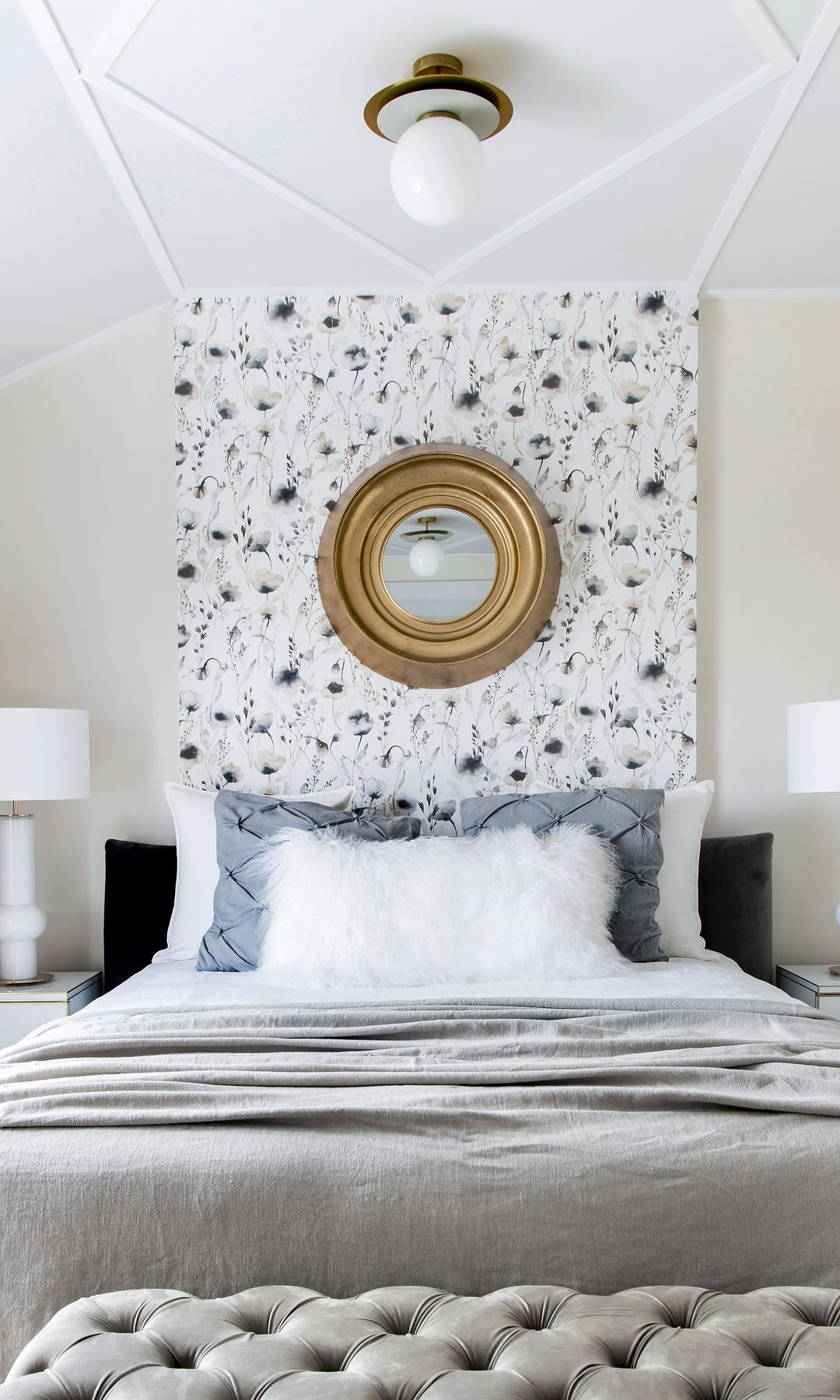 Dormitorio con pared del cabecero decorada, espejo redondo, lámparas de sobremesa y de techo, banqueta descalzado tapizada gris