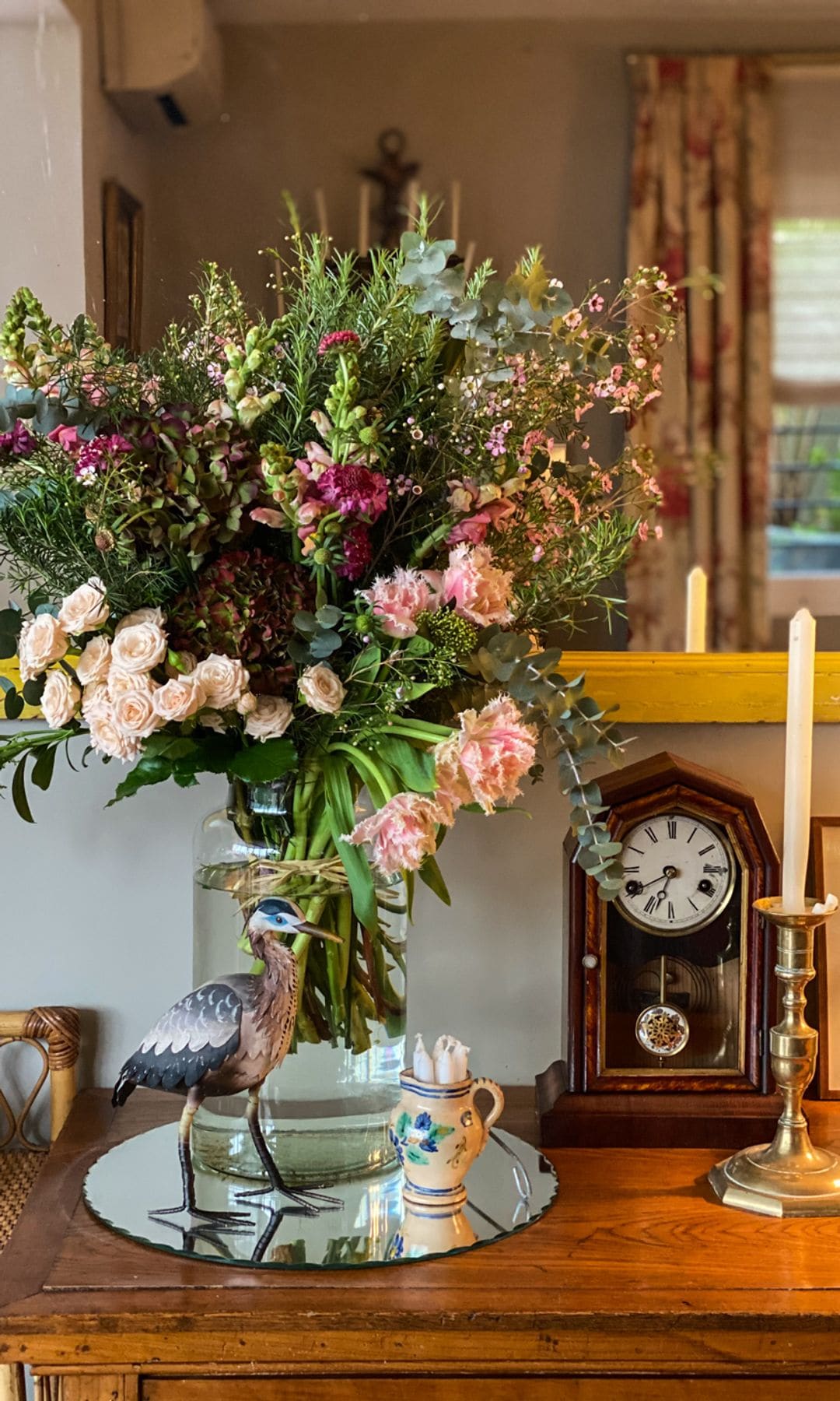 Jarrón de cristal transparente con flores en tonos rosas y verde, apoyado en un aparador, con candelabro dorado y reloj de madera de sobremesa