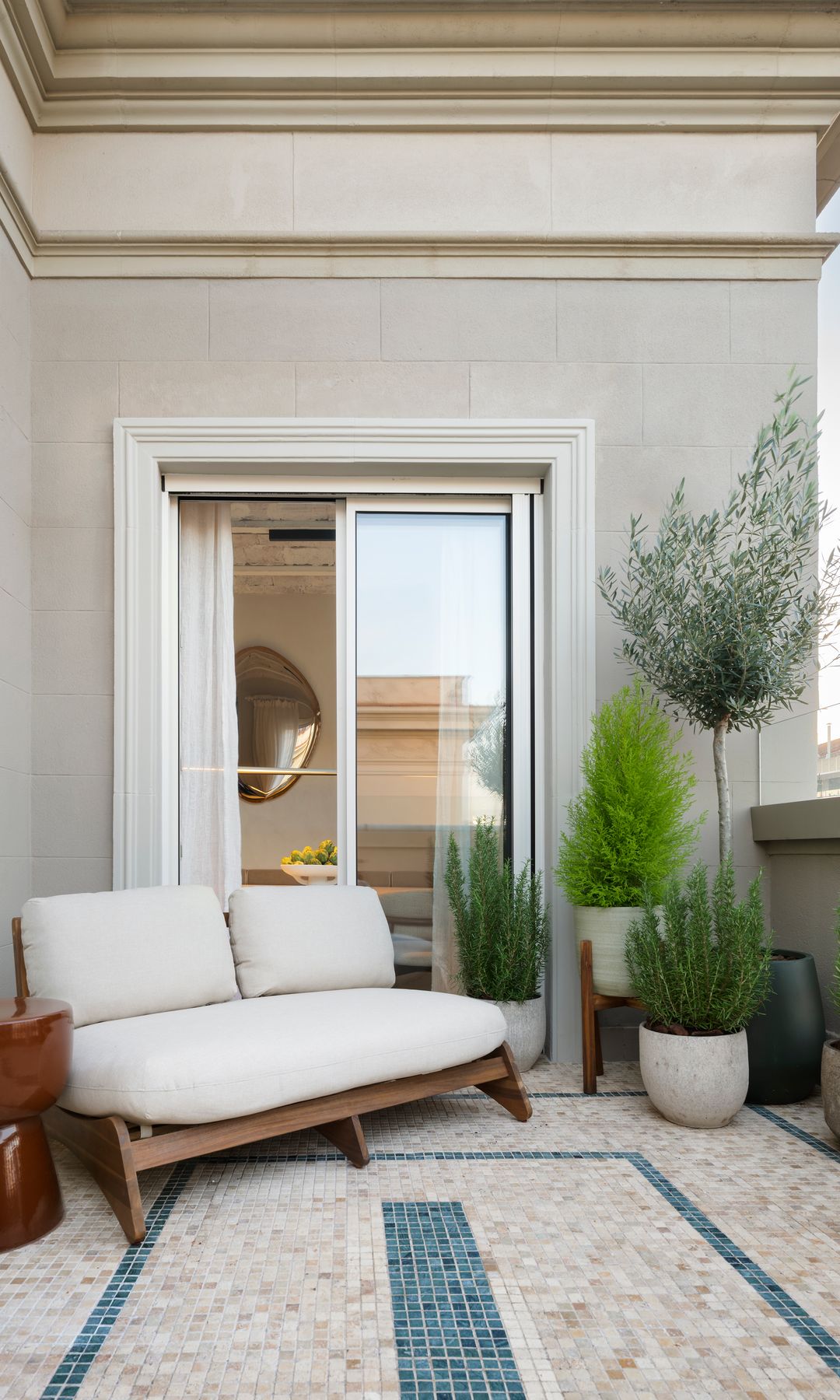 Terraza con suelo porcelánico, sofá de madera con cojines blancos, plantas verdes en maceta, olivo