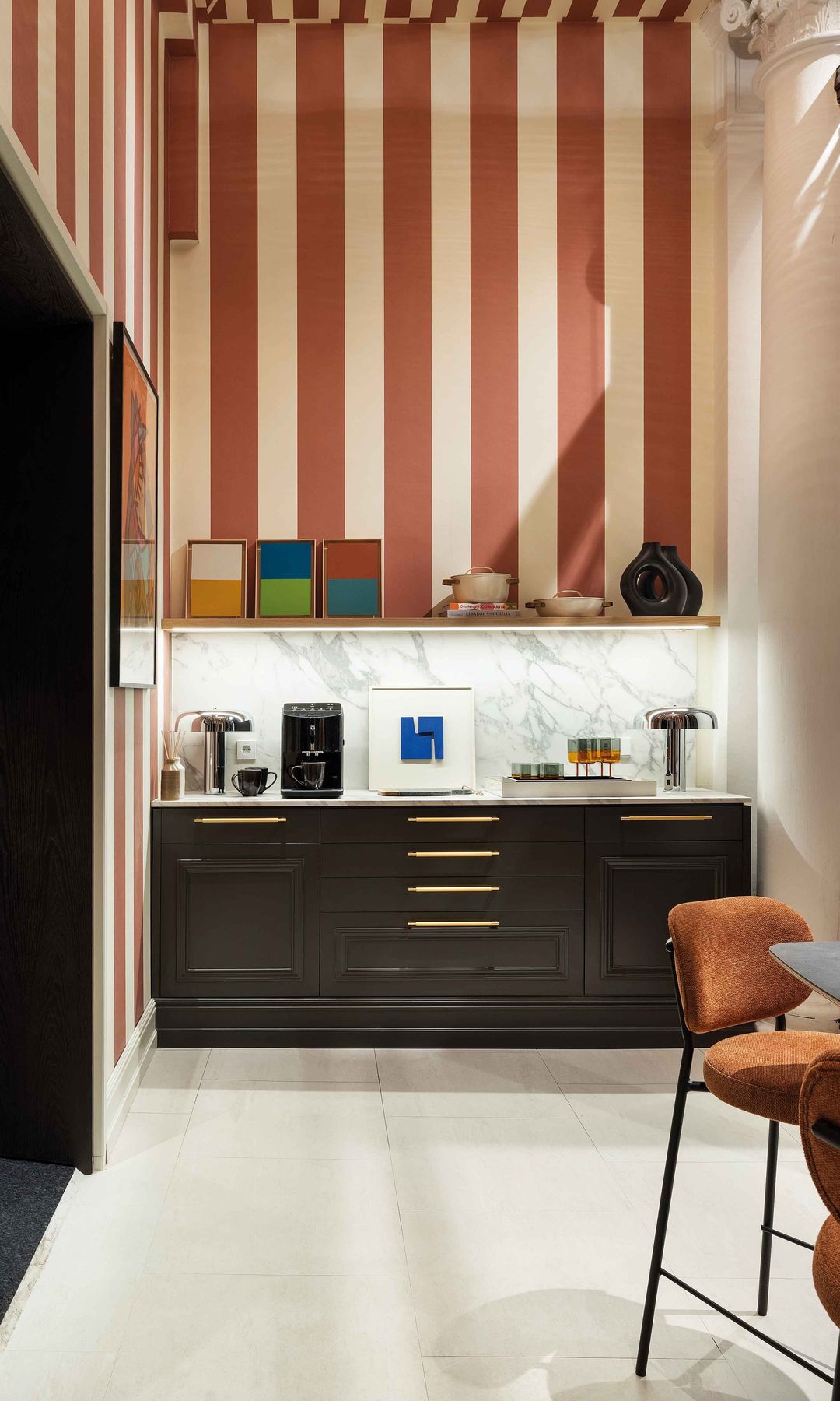 Rincón con papel pintado de rayas burdeos y blancas, mueble de desayuno con cafetera, tazas y cuadro, mesa con sillas