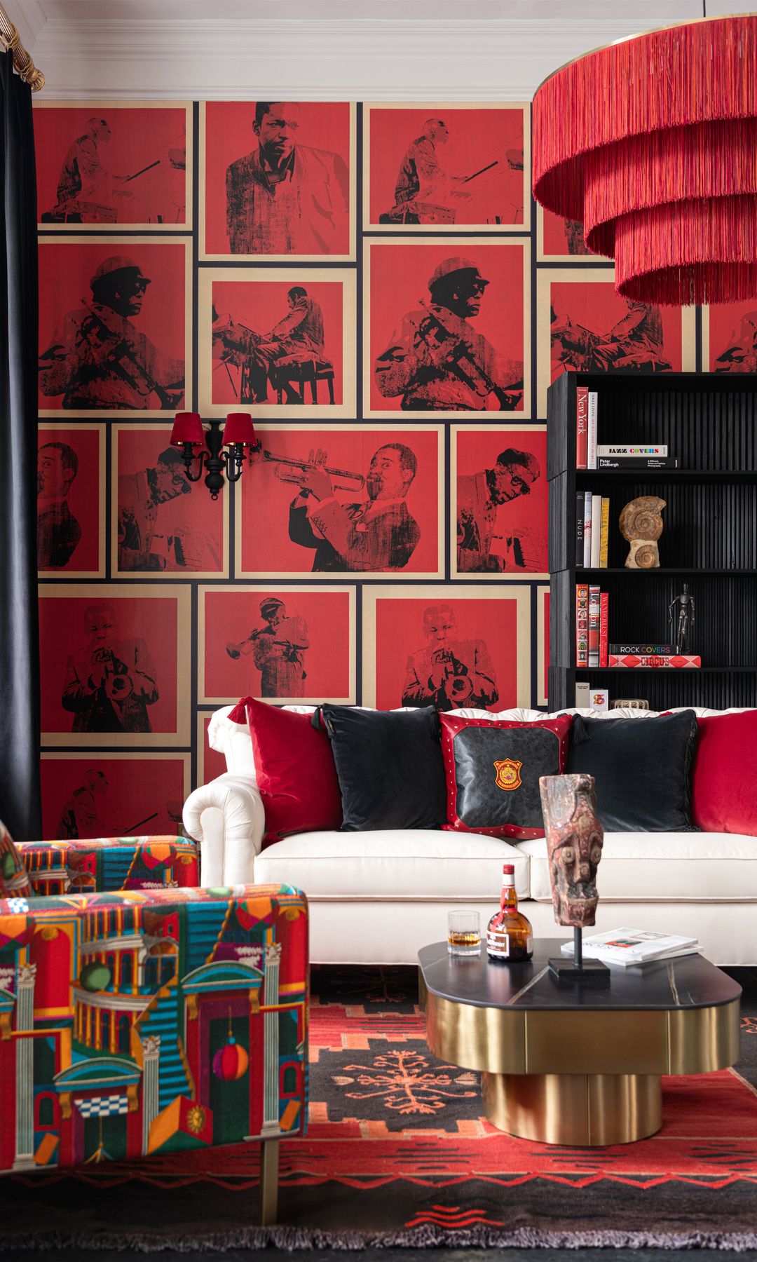 Salón con papel pintado de color rojo con figuras de jazz, sofá blanco, alfombra roja, butaca tapizada en colores, lámpara de techo de flecos rojas, cojines negros y rojos