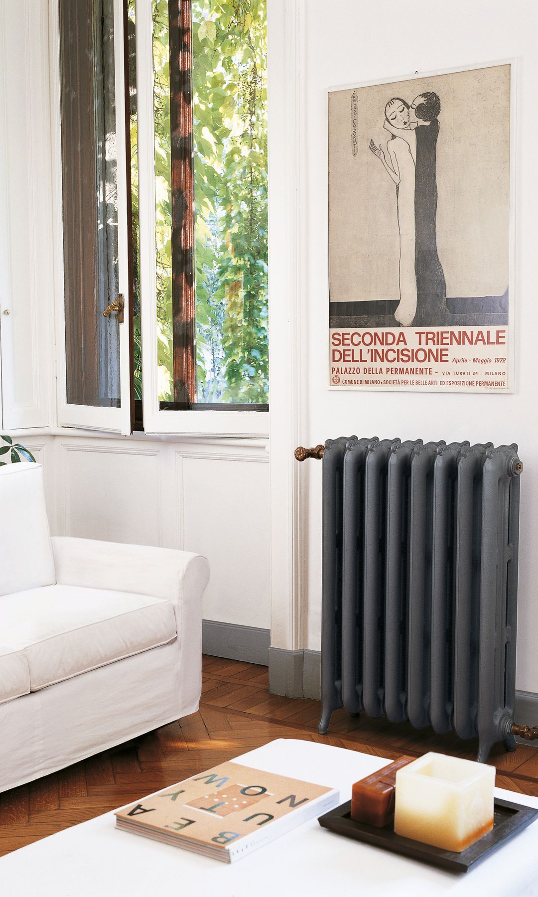 Salón con ventana, radiador antiguo en gris antracita, zócalo gris, sofá blanco, mesa de centro blanca, suelo de madera, póster en la pared