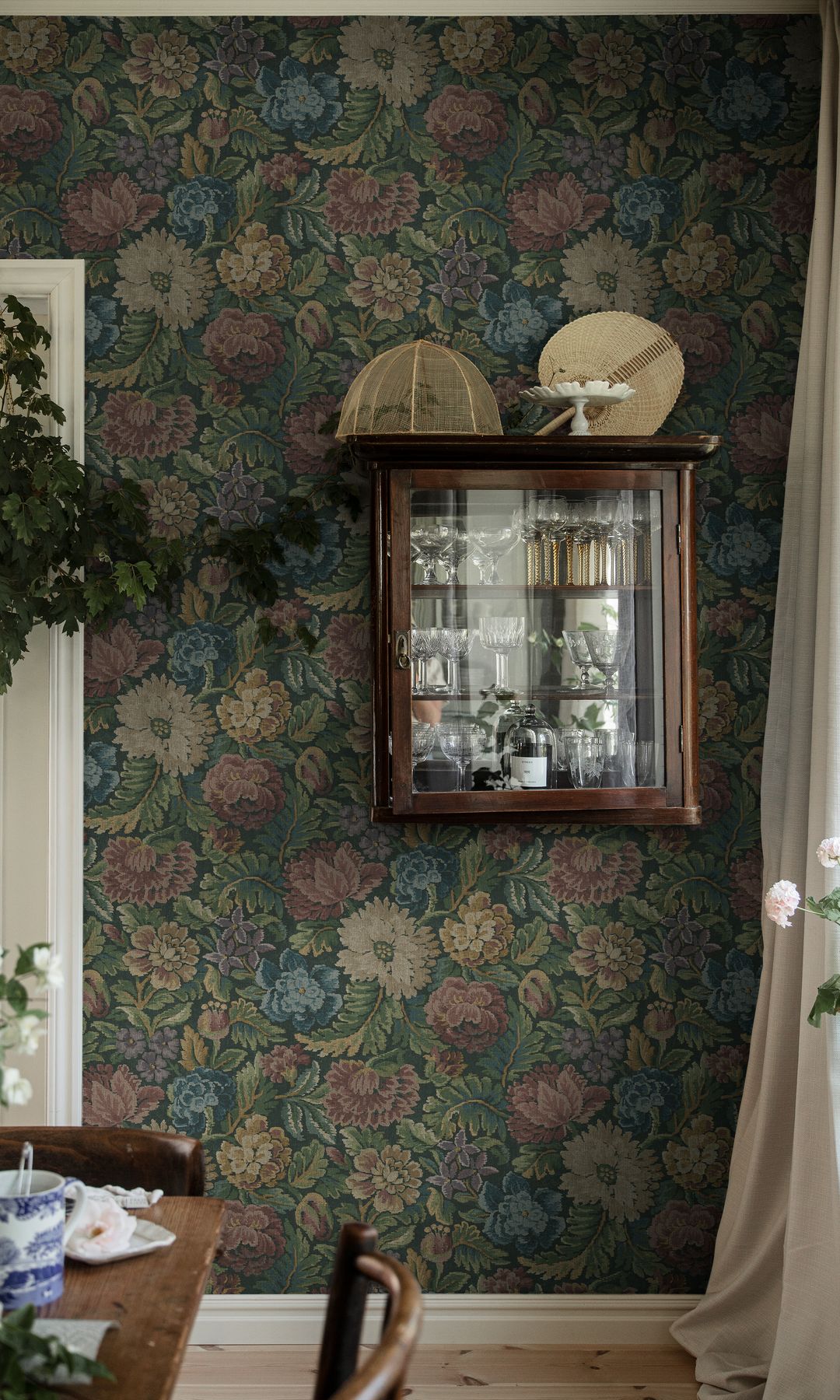 Comedor con mesa de madera, papel pintado de flores, vitrina en la pared de madera con vajillas y platos 