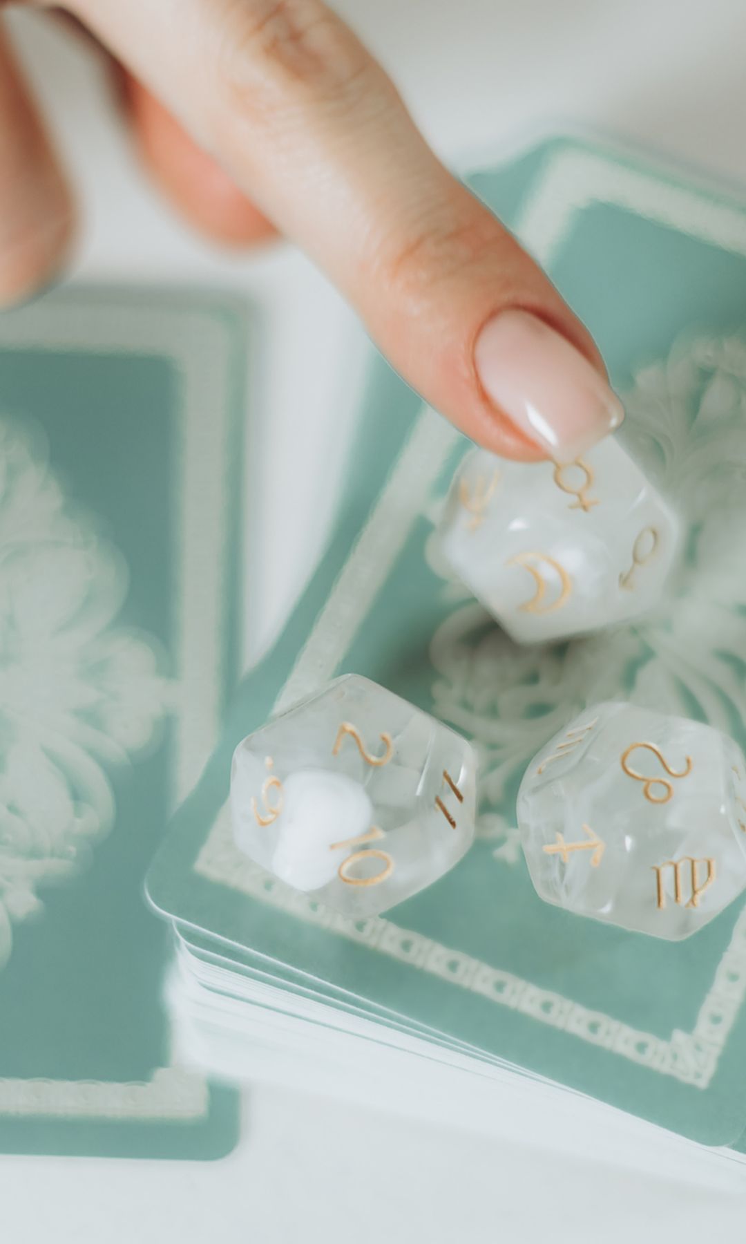 Primer plano de un dedo de mujer señalando un dado de juego de 12 caras que descansa sobre una baraja de cartas colocada sobre una superficie blanca.