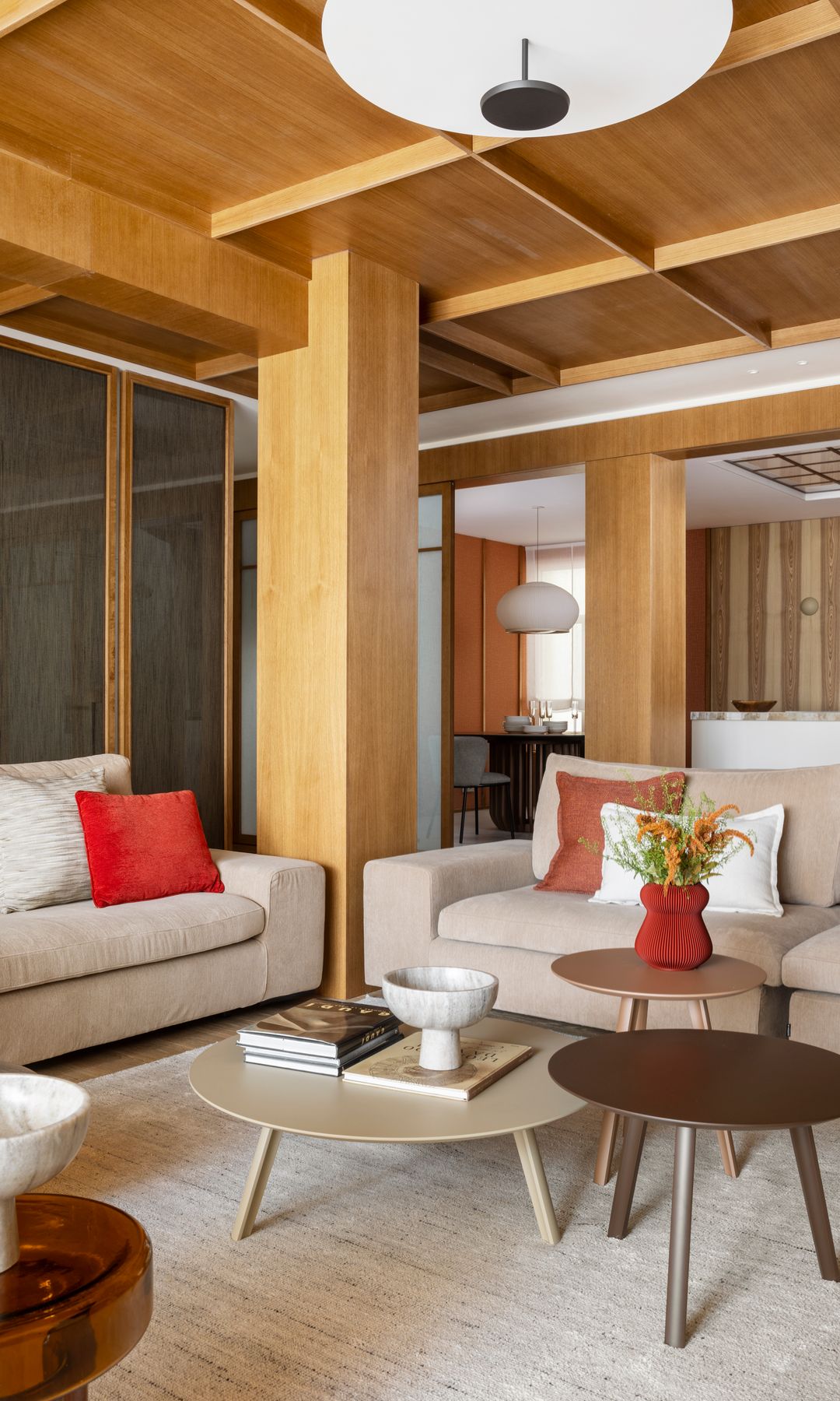 Salón con techo y paredes de madera, sofás blancos, mesas de centro, cojines rojos y florero rojo