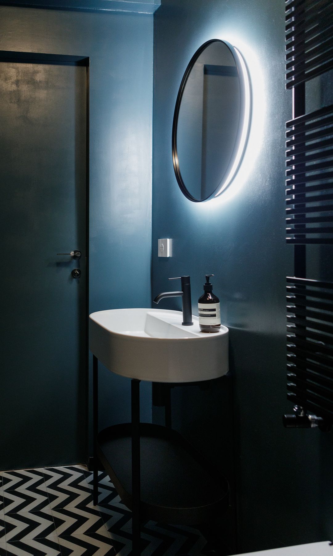 Aseo con paredes en color azul intenso, espejo redondo retroiluminado, suelo en blanco y negro, radiador toallero, lavabo negro con pie negro 