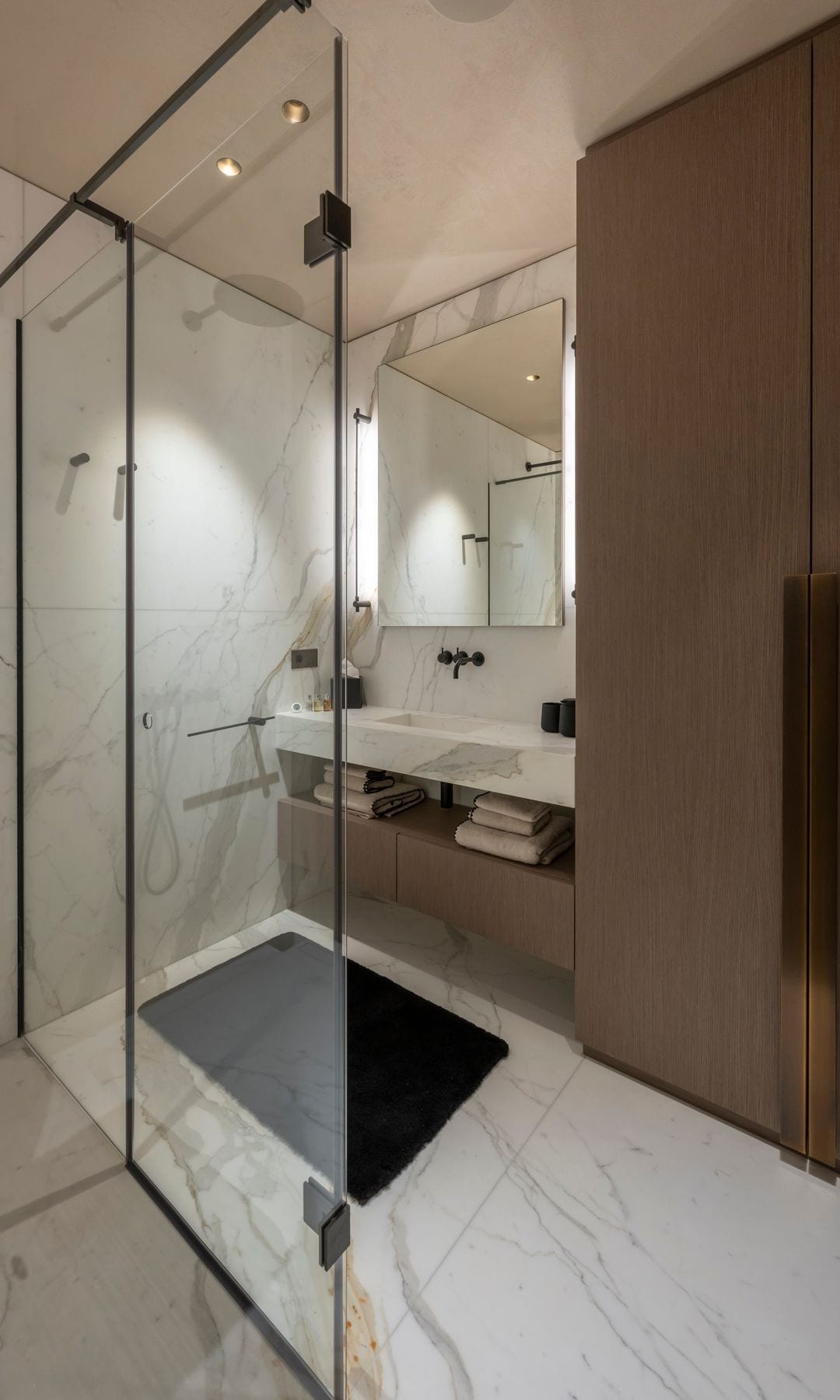 Baño con suelo y paredes de mármol, mampara de ducha, armario de madera, espejo
