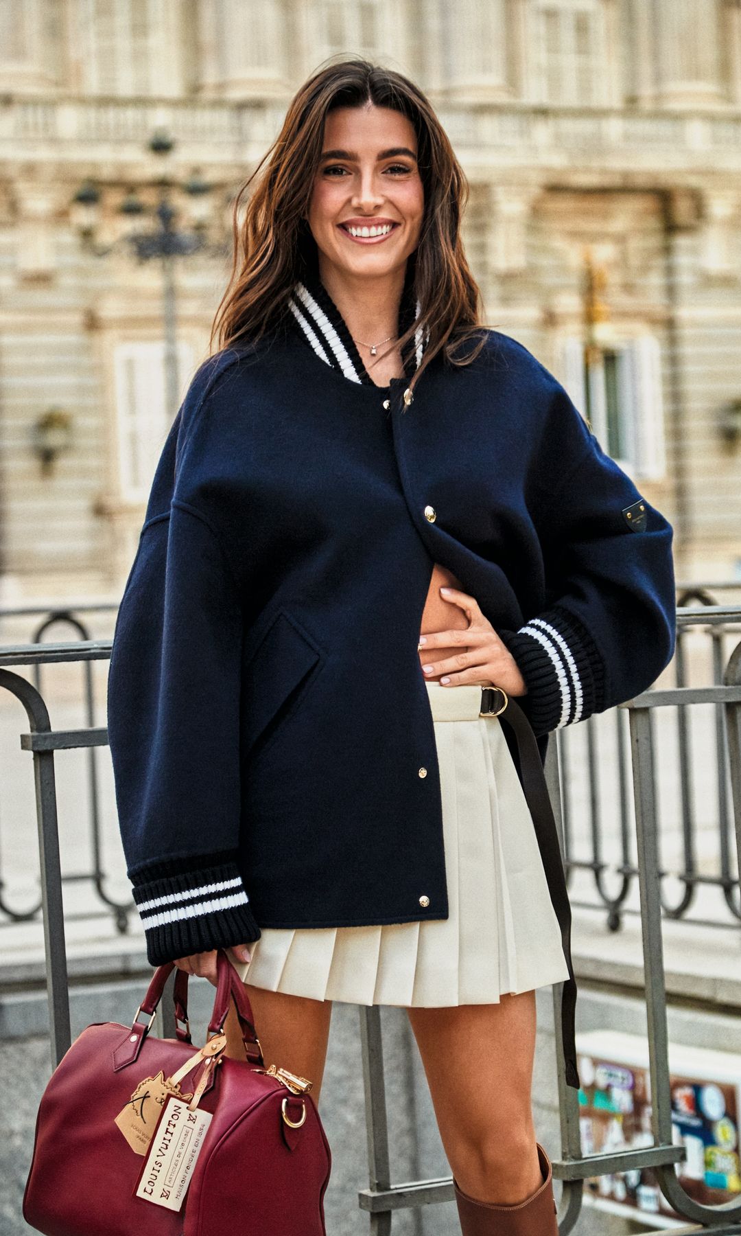 Gemma con chaqueta universitaria de doble cara, minifalda plisada con cinturón, confeccionada en gabardina técnica, y bolso Speedy Soft 30 actualizado para la colección LV Travels with Grace Coddington, todo de Louis Vuitton