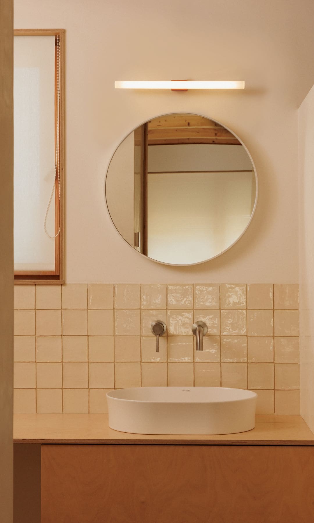 Baño con azulejos en la pared del lavabo cuadrados en color beige, espejo redondo, aplique de pared, lavabo sobre la encimera 