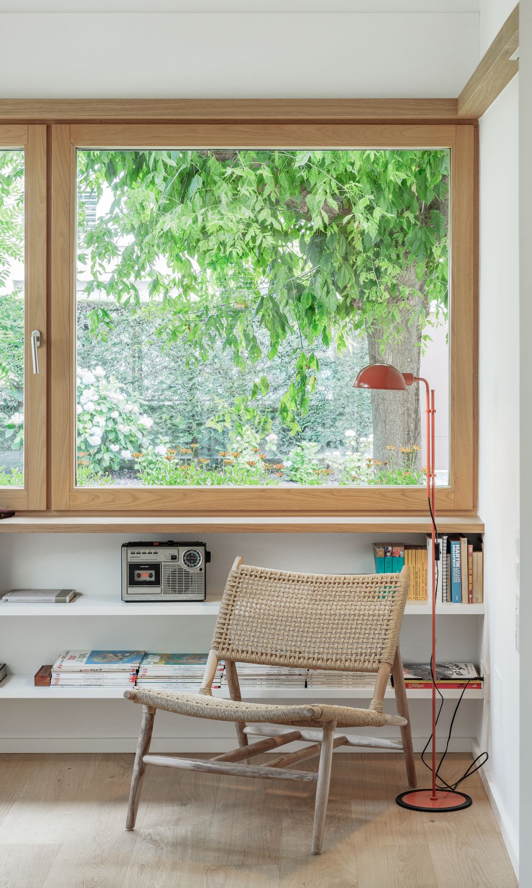 Rincón de lectura con estantería bajo la ventana, silla de enea, lámpara de pie roja 
