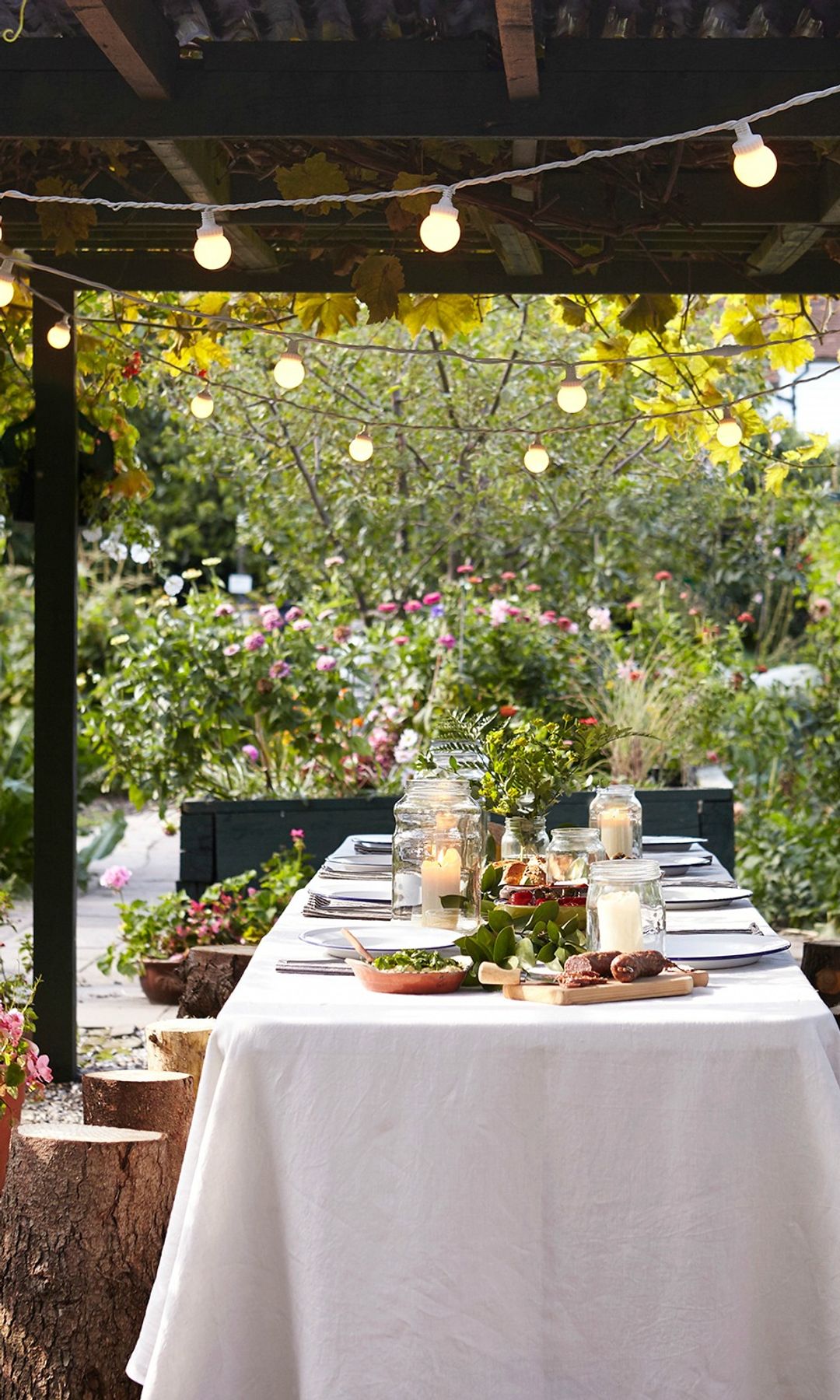 Mesa bajo pérgola de madera con mantel blanco, guirnalda de luces, troncos de árbol como asiento, macetas y flores 