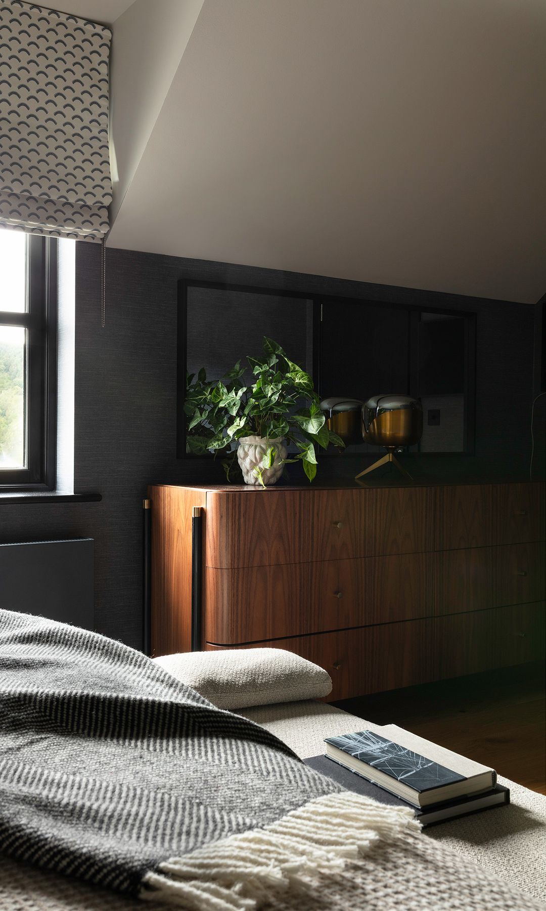 Dormitorio con paredes grises, estor, ropa de cama gris claro, techo abuhardillado crema, aparador de madera con plantas encima 