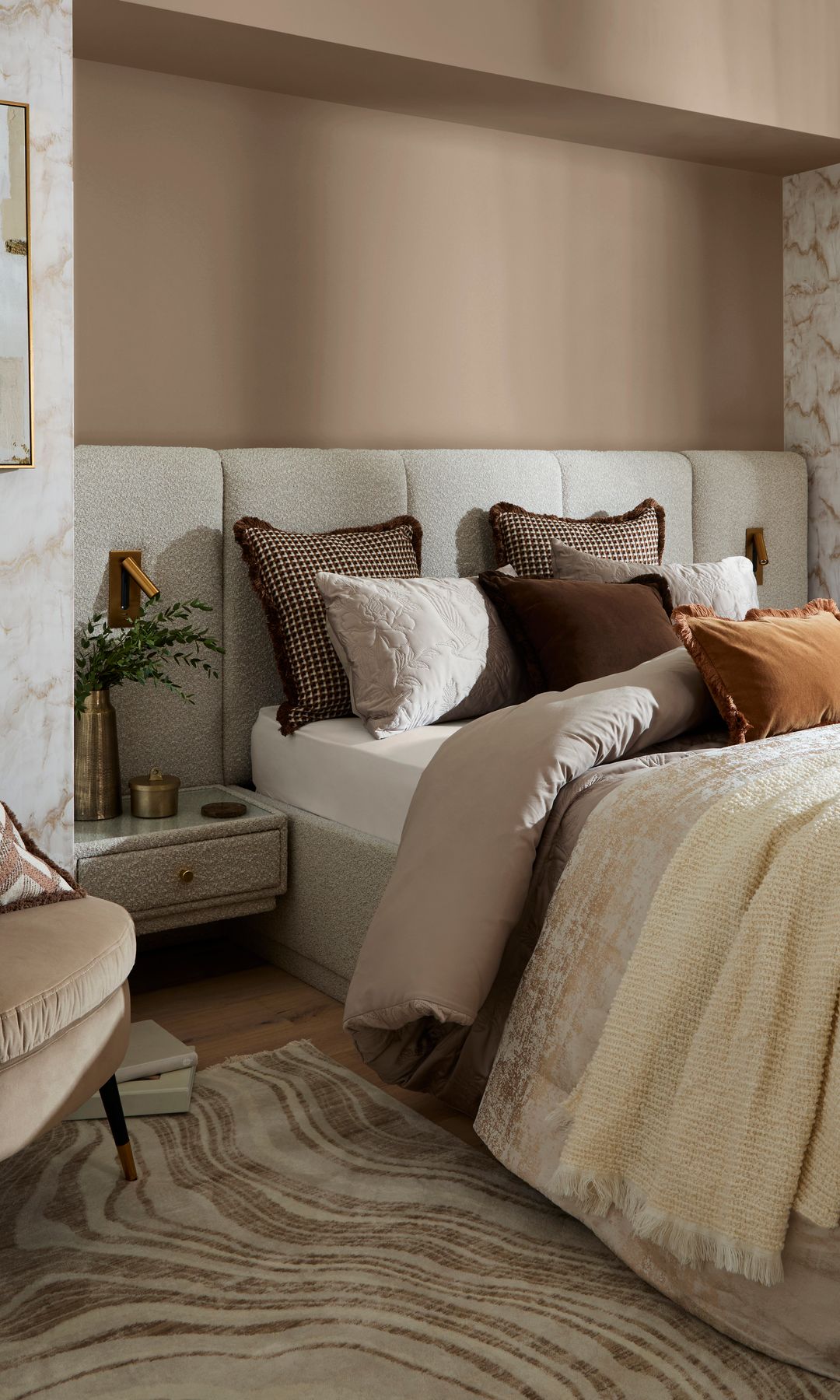 Dormitorio con cabecero tapizado de pared a pared en lana bouclé, alfombra, cojines en tonos tierra, mesita de noche de lana bouclé, jarrón dorado, butaca de terciopelo beige