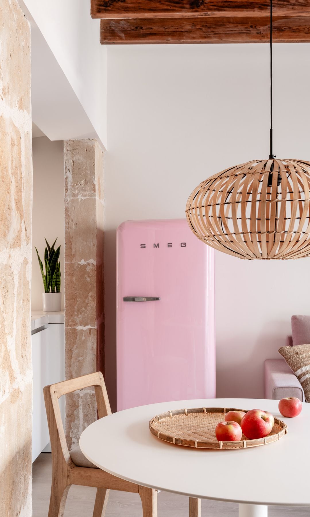 Comedor con nevera Smeg de color rosa, mesa blanca, lámpara de techo, vigas de madera en el techo y frutero con manzanas