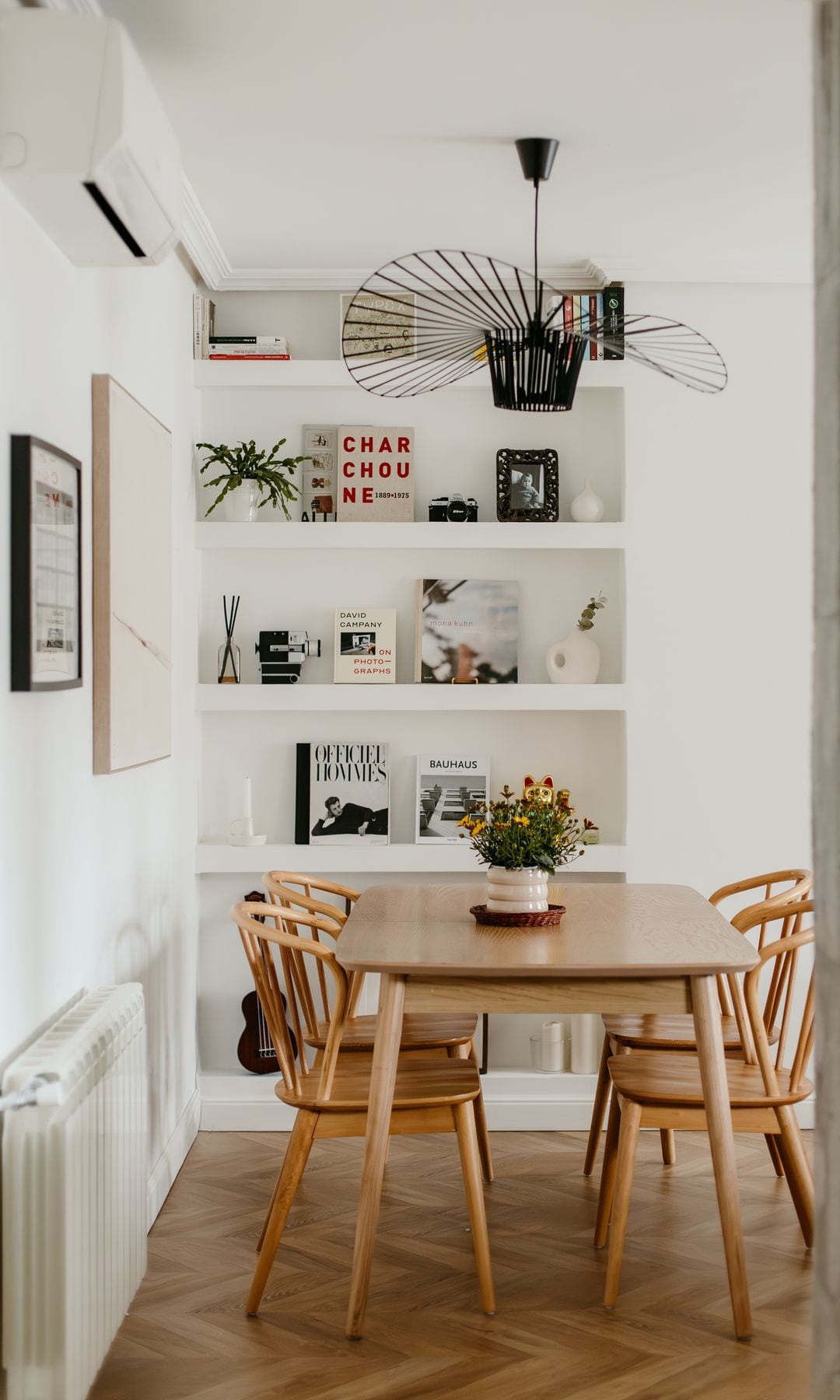 Comedor con mesa de madera, sillas de madera, lámpara de techo negra, maceta blanca con flores sobre la mesa, cuadros en la pared, estantería de obra con libros y adornos