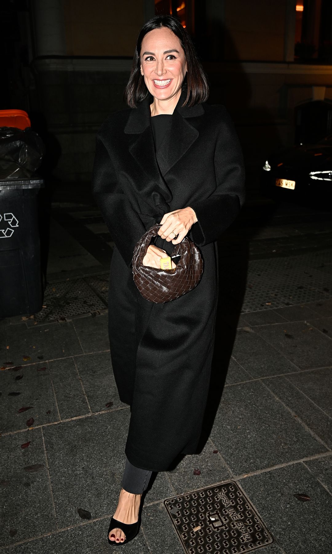 Tamara Falcó  con abrigo negro largo, sonriendo y sosteniendo un bolso marrón, caminando en la calle.
