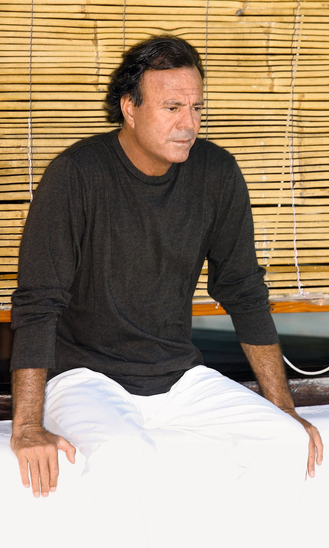 Julio Iglesias sentado con expresión serena, vestido con camiseta y pantalones blancos.