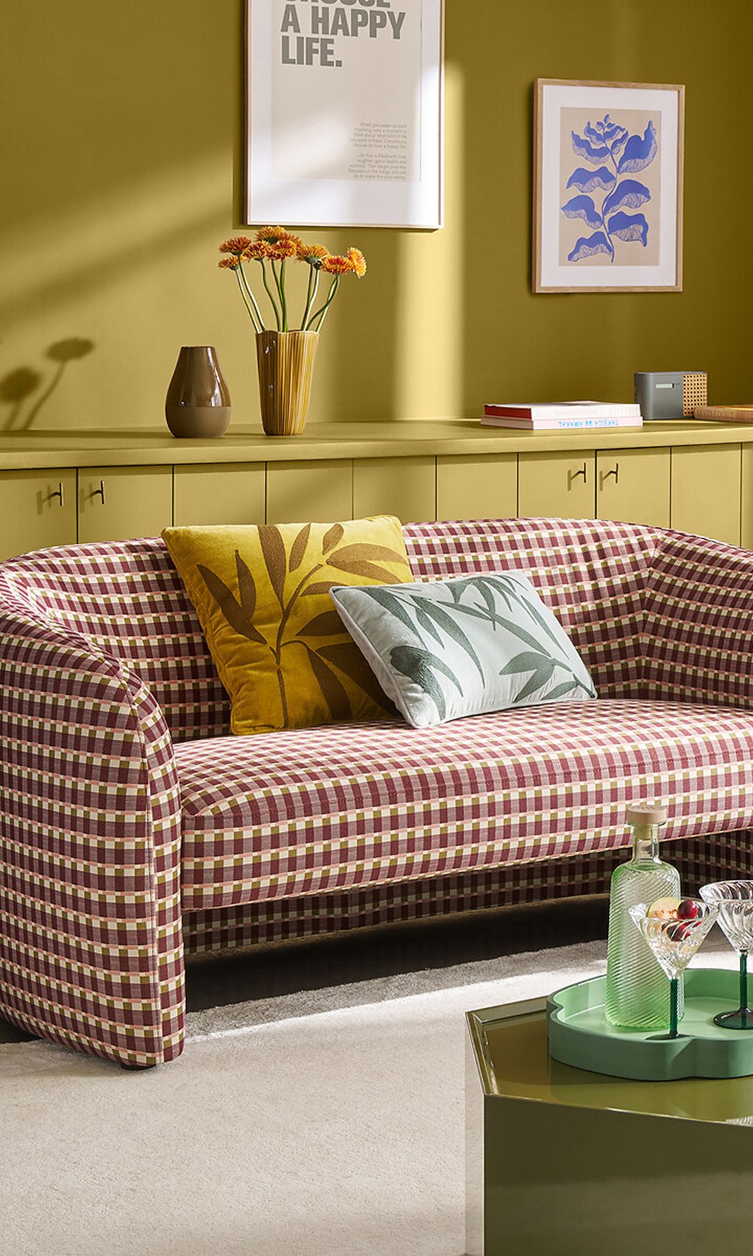 Salón con sofá de cuadros burdeos y blancos, mueble y paredes de color verde oliva, mesa de centro verde pino y verde oliva, cojines con estampados vegetales en amarillo y verde 