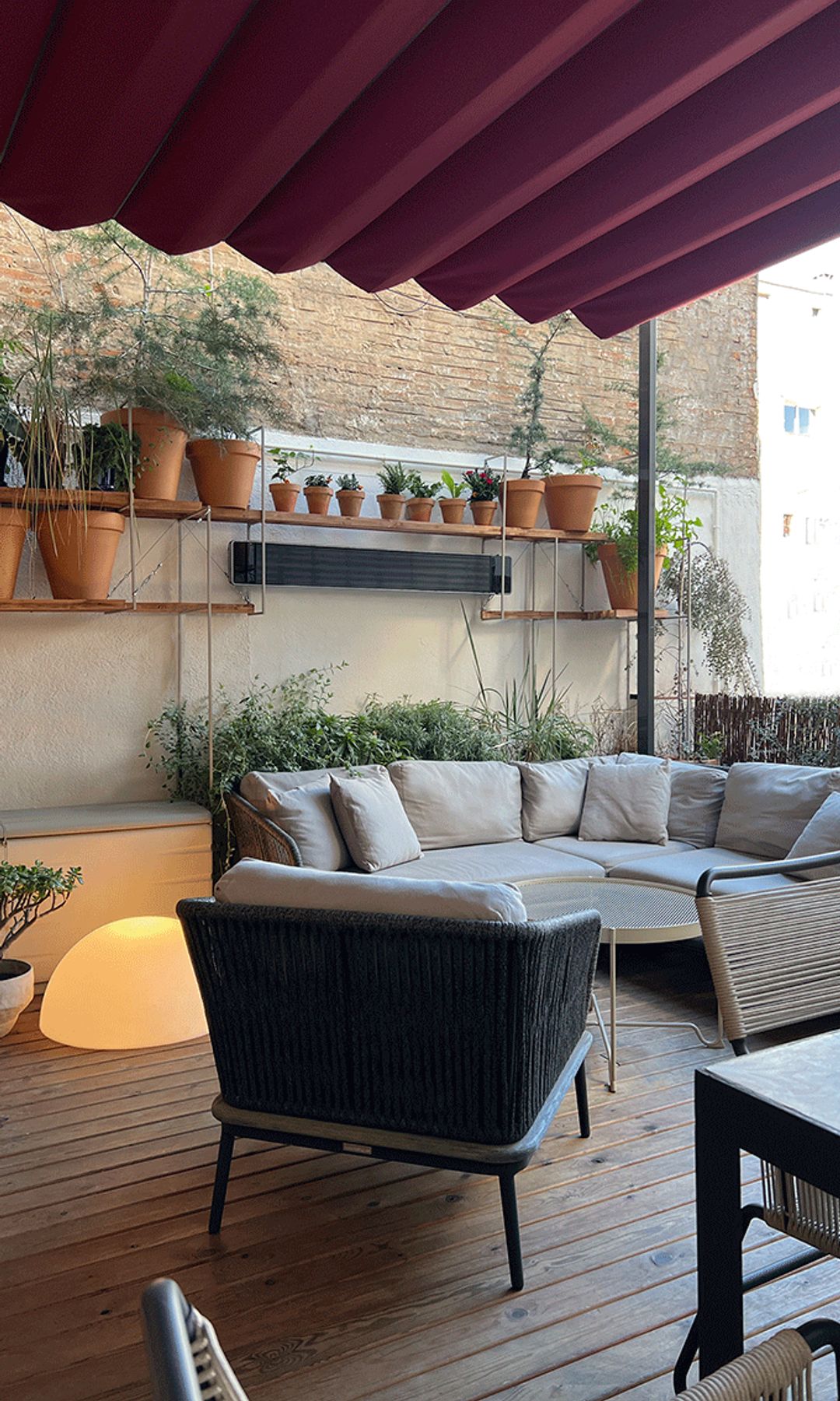 Terraza urbana con pérgola roja, suelo de madera, sofá, baldas de madera con macetas con plantas, sofá gris 