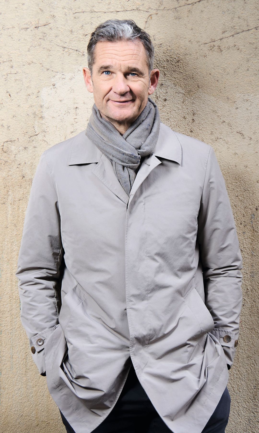 Iñaki Urdangarin sonriendo, con abrigo gris y bufanda, contra pared texturizada.