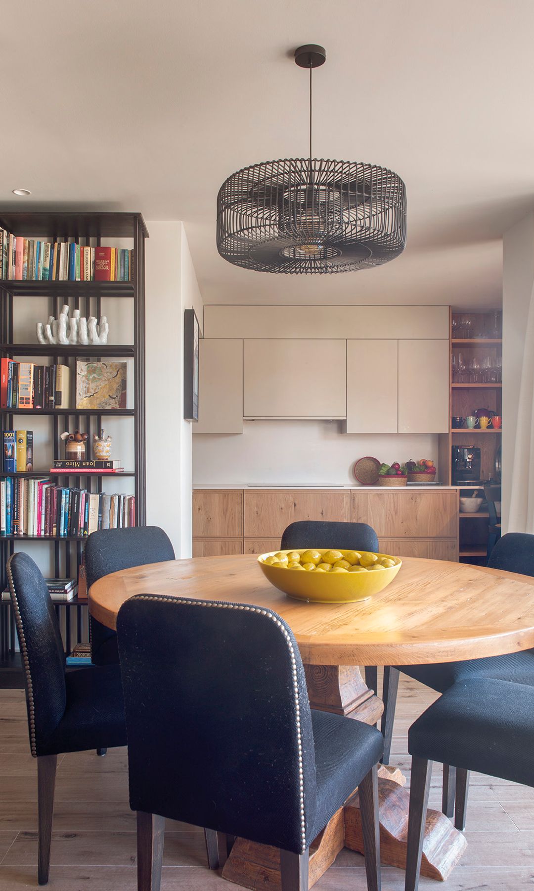 Salón comedor y cocina, con estantería negra, mesa redonda de comedor, sillas tapizadas, muebles de cocina en blanco y madera 