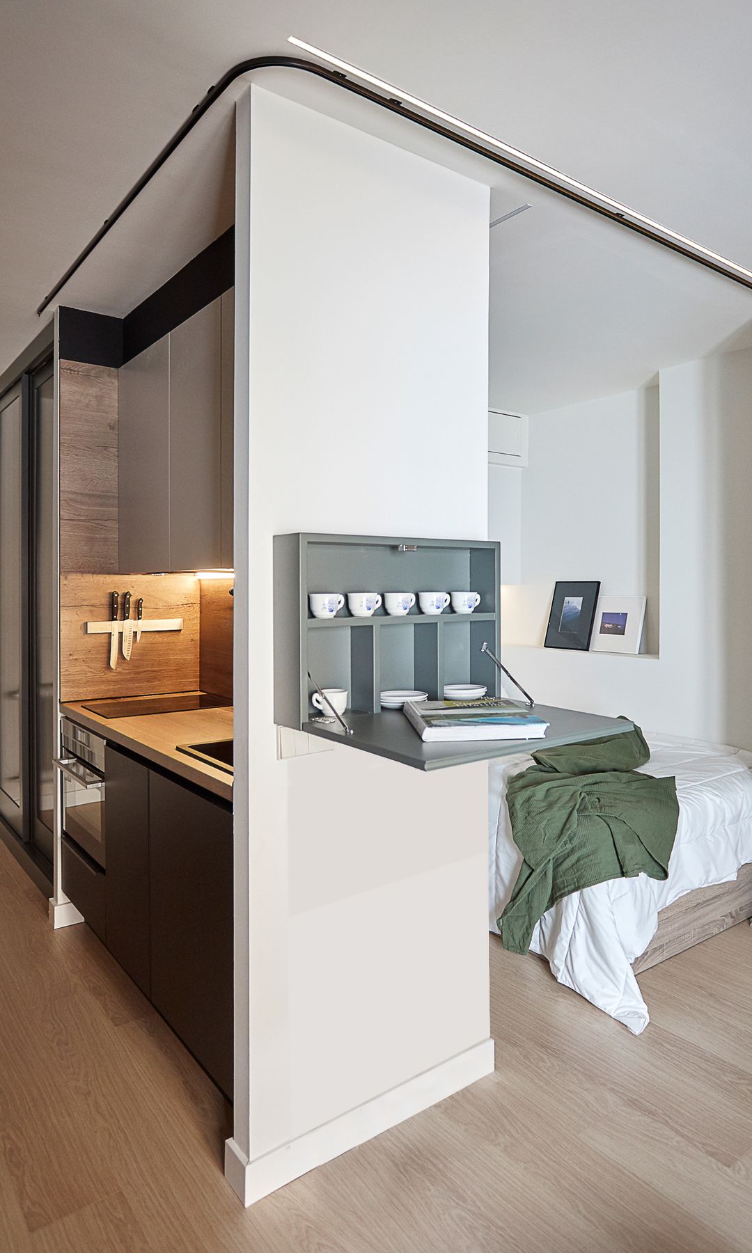 Piso pequeño con estructura que alberga cama en un lado y cocina por el otro, con repisa con platos y tazas colgada en la pared plegable