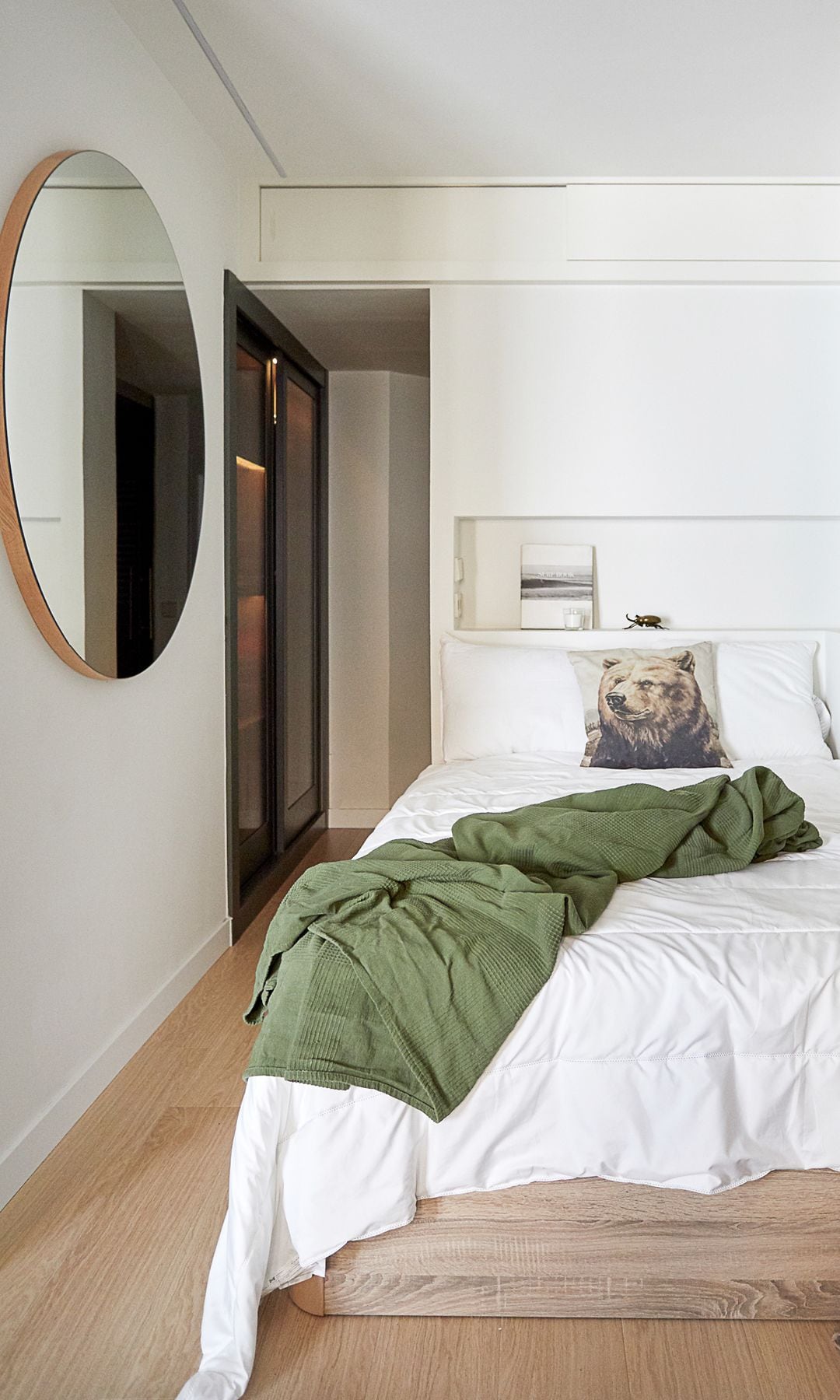Dormitorio con pasillo con zona de armarios, espejo redondo, suelo de madera, ropa de cama blanca, colcha verde, hornacina en la pared del cabecero, cojín con la imagen de un oso en la cama