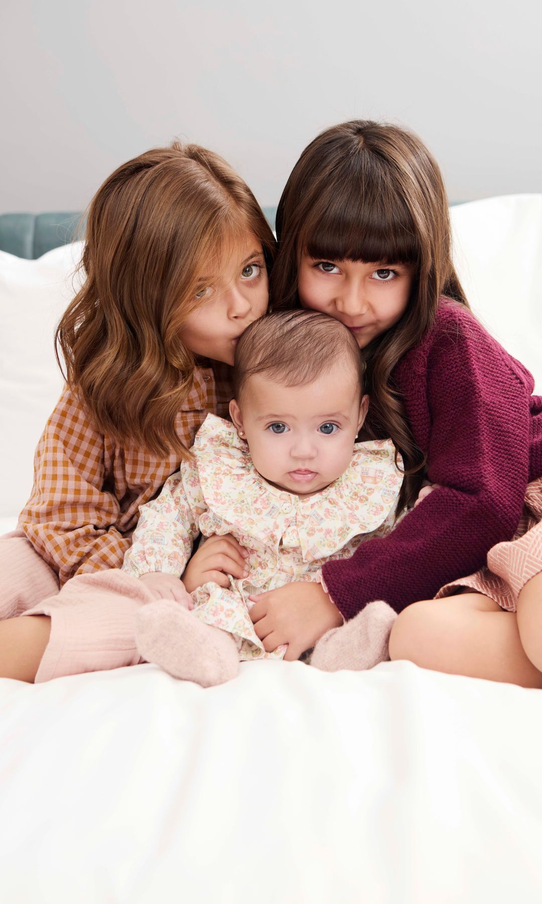 Tres niñas en una cama, las mayores besan a la bebé en la cabeza