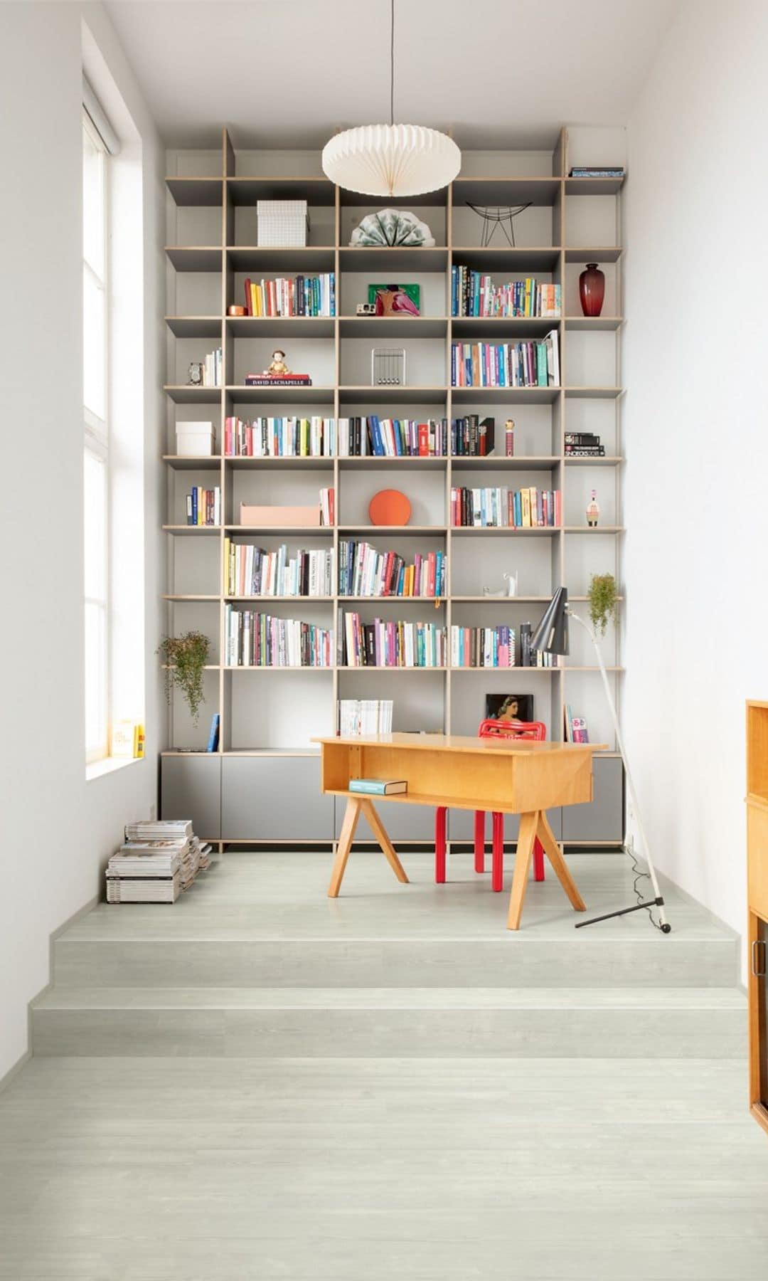 Despacho con librería de suelo a techo, mesa de madera, dos escalones, suelo vinílico blanco, lámpara de techo 