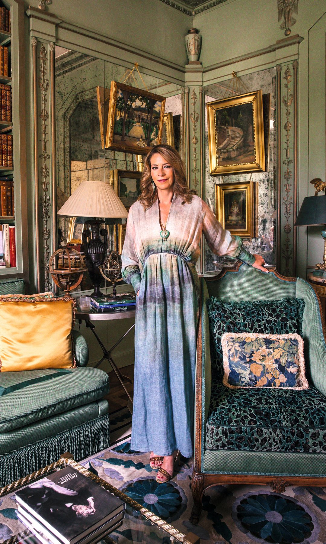 La baronesa Jacqueline von Hammerstein-Loxten, en vestido azul y sandalias, de pie en una sala elegante con muebles verdes y libros.