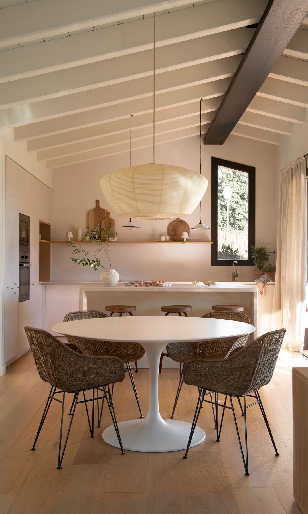Cocina blanca abierta al comedor, con techo de madera blanco, mesa de comedor redonda, sillas de fibras vegetales, lámpara de techo de papel