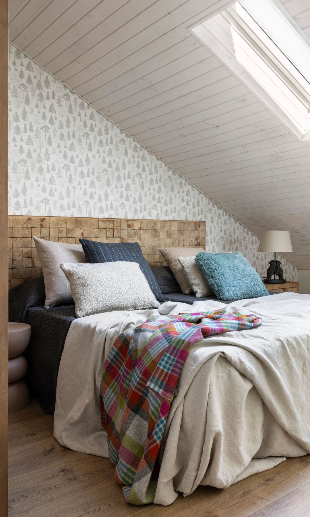 Dormitorio abuhardillado con techo de lamas de madera blancas, papel pintado en la pared del cabecero, cabecero de madera, cojines, ventana techo