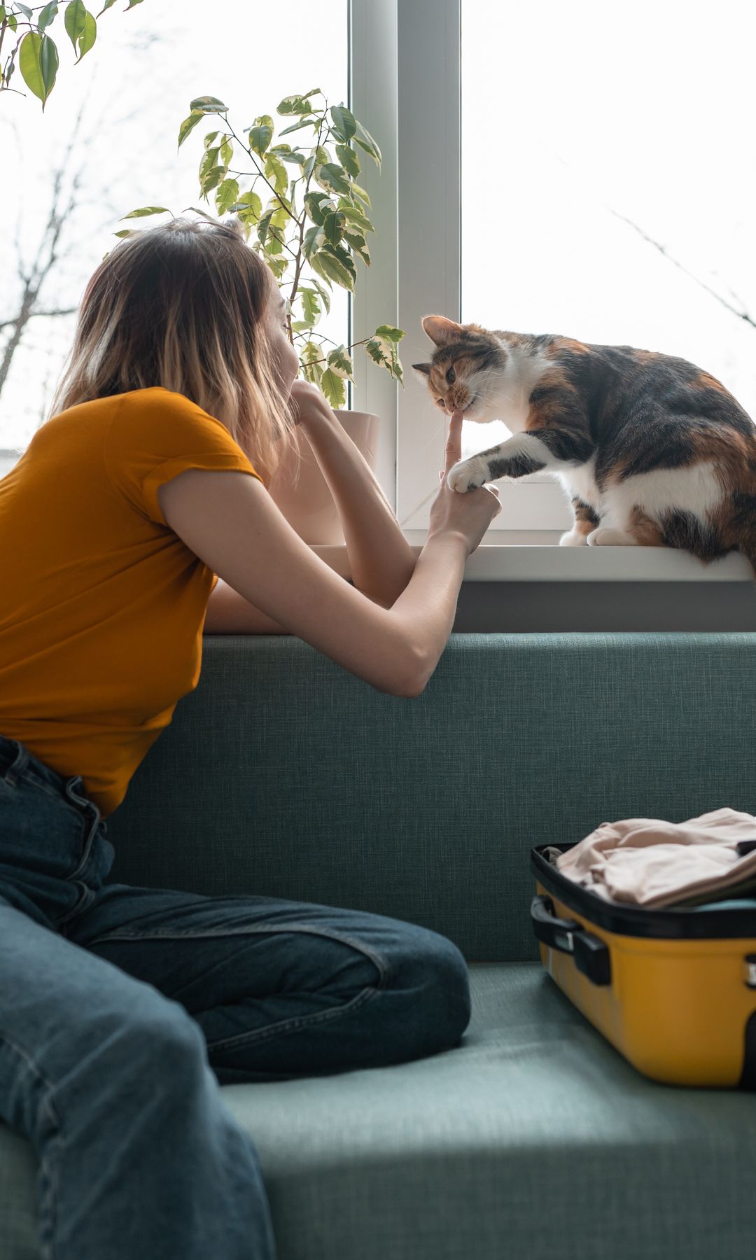 mujer con un gato junto a la ventana, con una maleta abierta sobre el sofá