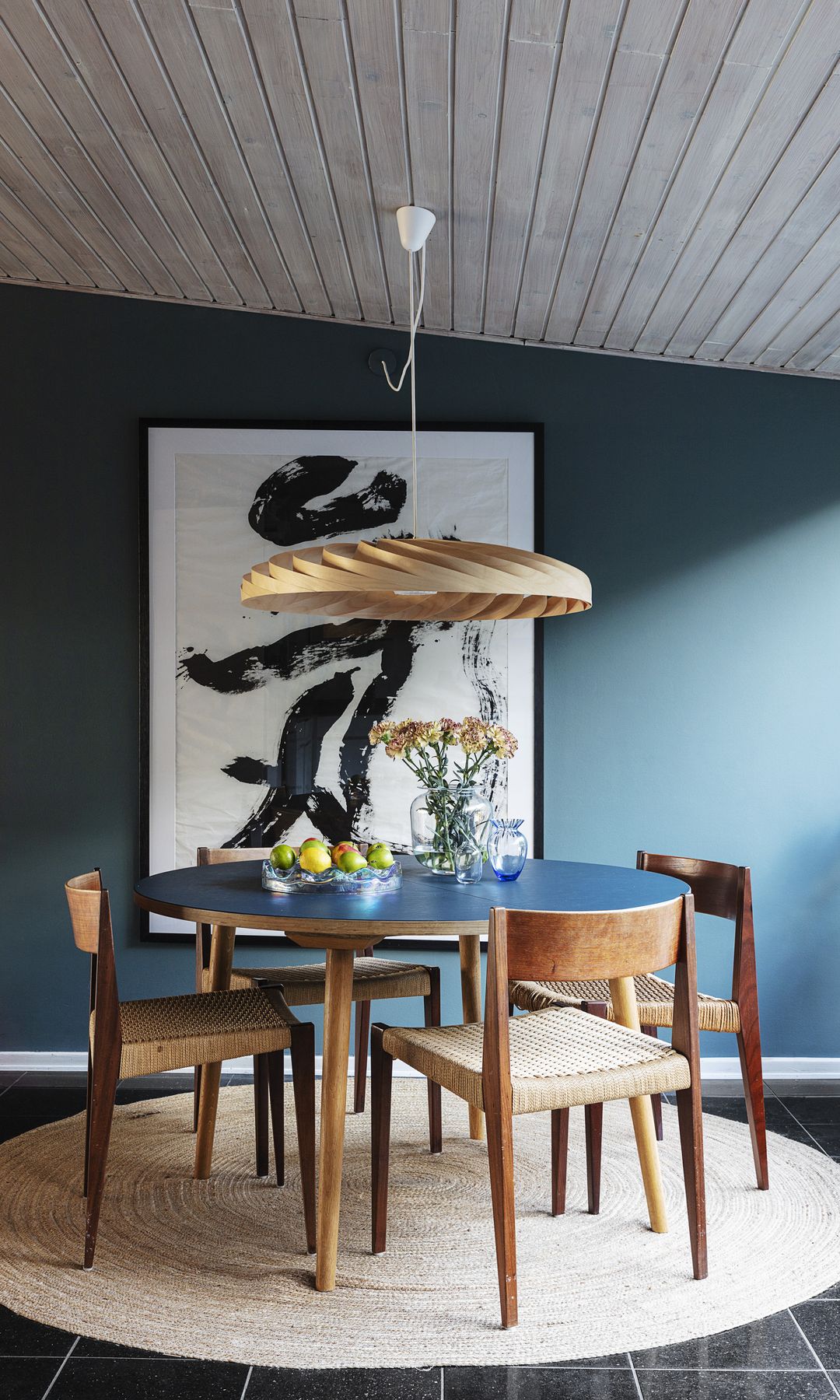 Comedor con mesa redonda, paredes de color azul petróleo, sillas de madera, cuadro en la pared, lámpara de techo, suelo negro
