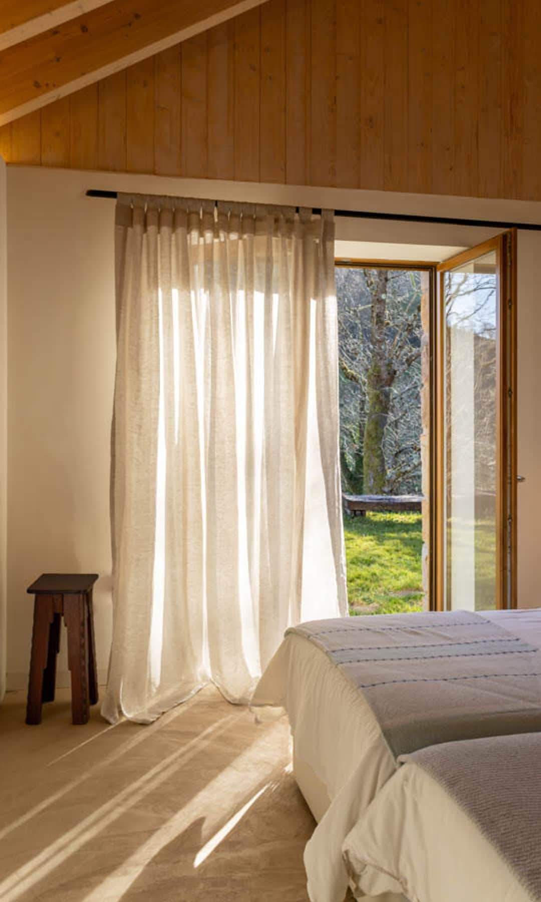 Dormitorio con salida al jardín con ventanas con cortina blanca de lino techo de madera, taburete de madera y dos camas