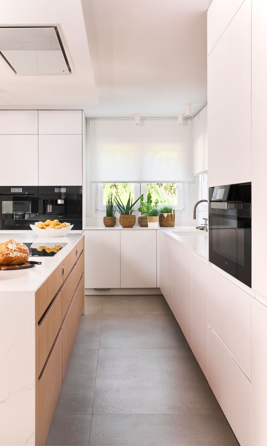 Cocina blanca con distribución paralela, isla de cocina, campana en el techo, muebles sin tiradores y estor blanco