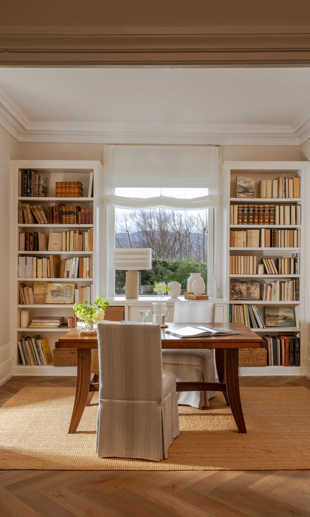 Despacho con mesa de madera, silla con funda, alfombra, librerías blancas a ambos lados de la ventana. estor blanco, lámpara de sobremesa 