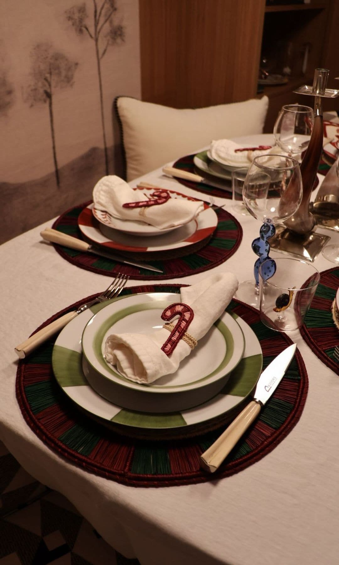 Mesa de Navidad con mantel blanco, manteles individuales burdeos, vajilla verde y blanca, servilletero rosa en forma de bastón, copas y vasos 